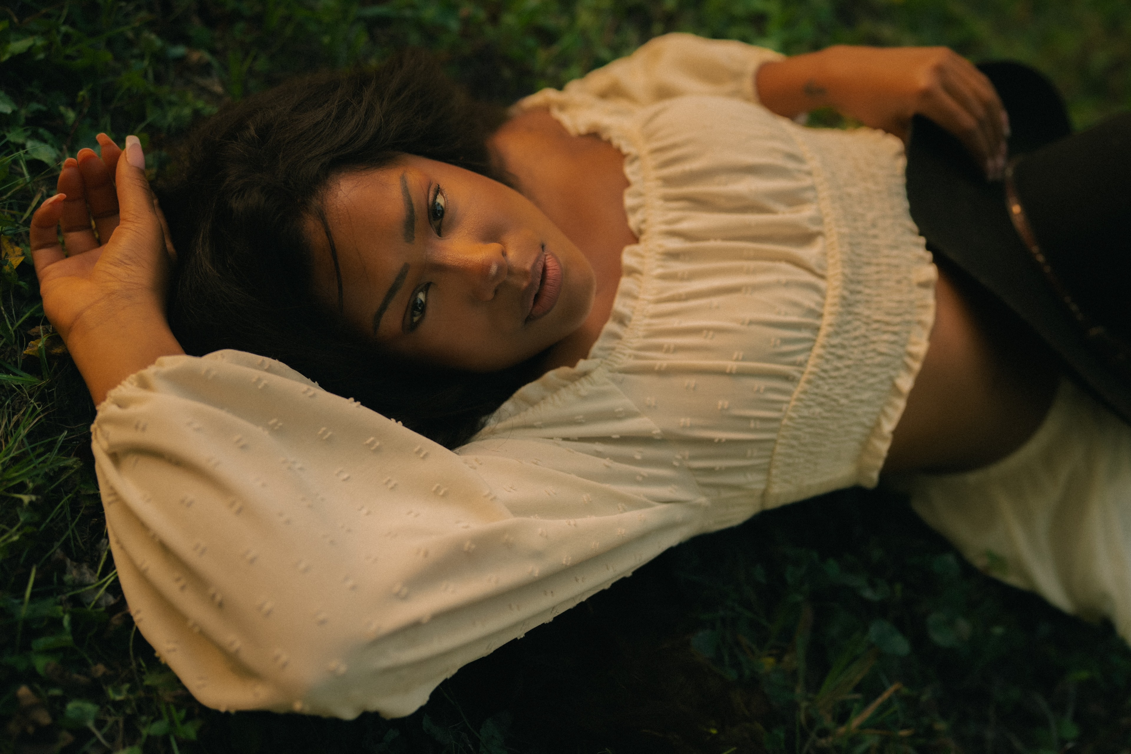 Woman in a cream smocked-bodice blouse lying in grass, head tilted toward camera, one hand raised near her hair, the other resting on a dark hat beside her, warm low-angled light on her face.