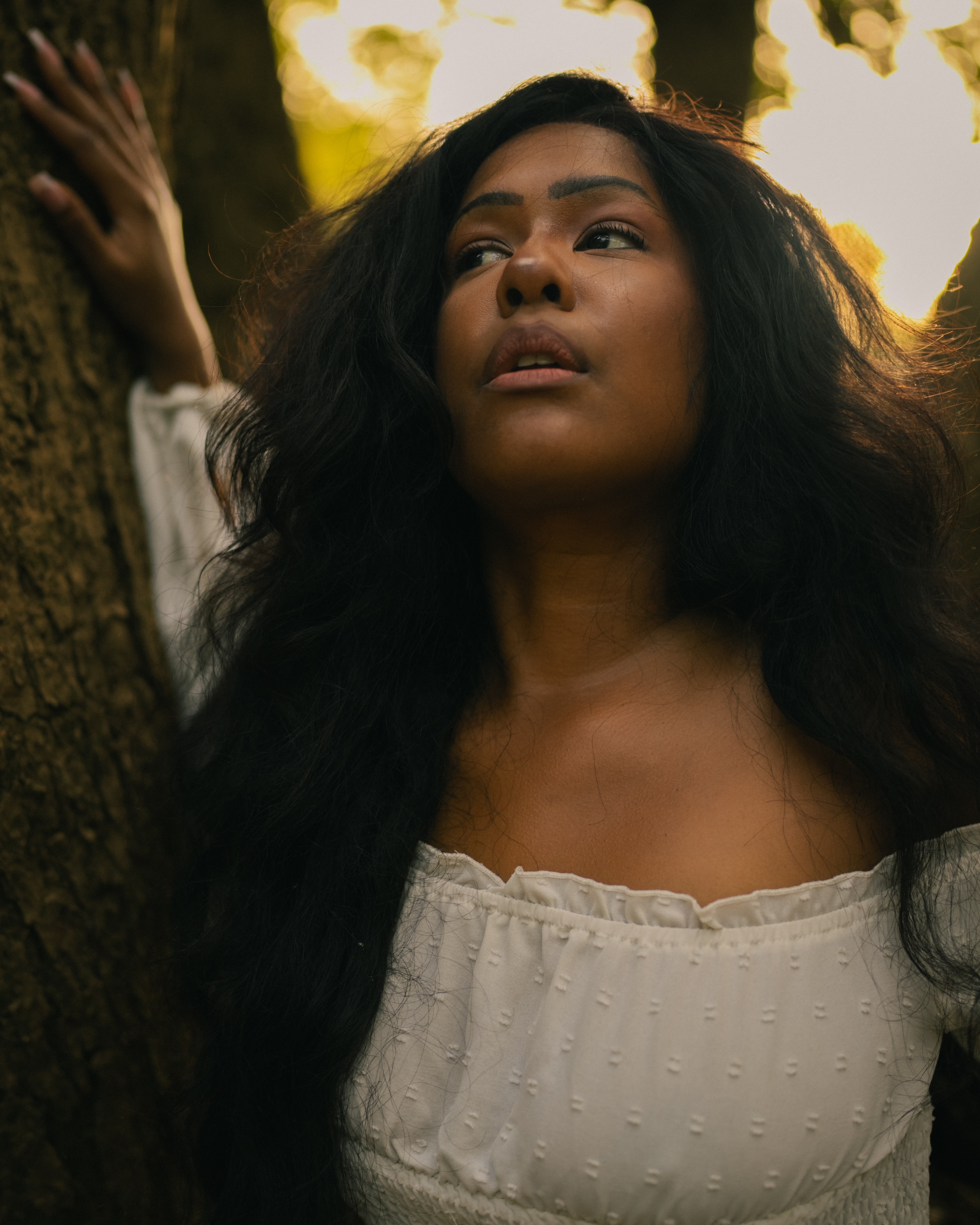 Woman in an off-shoulder white top rests one raised hand on tree bark, head tilted back and eyes glancing sideways in warm golden backlight filtering through leaves.