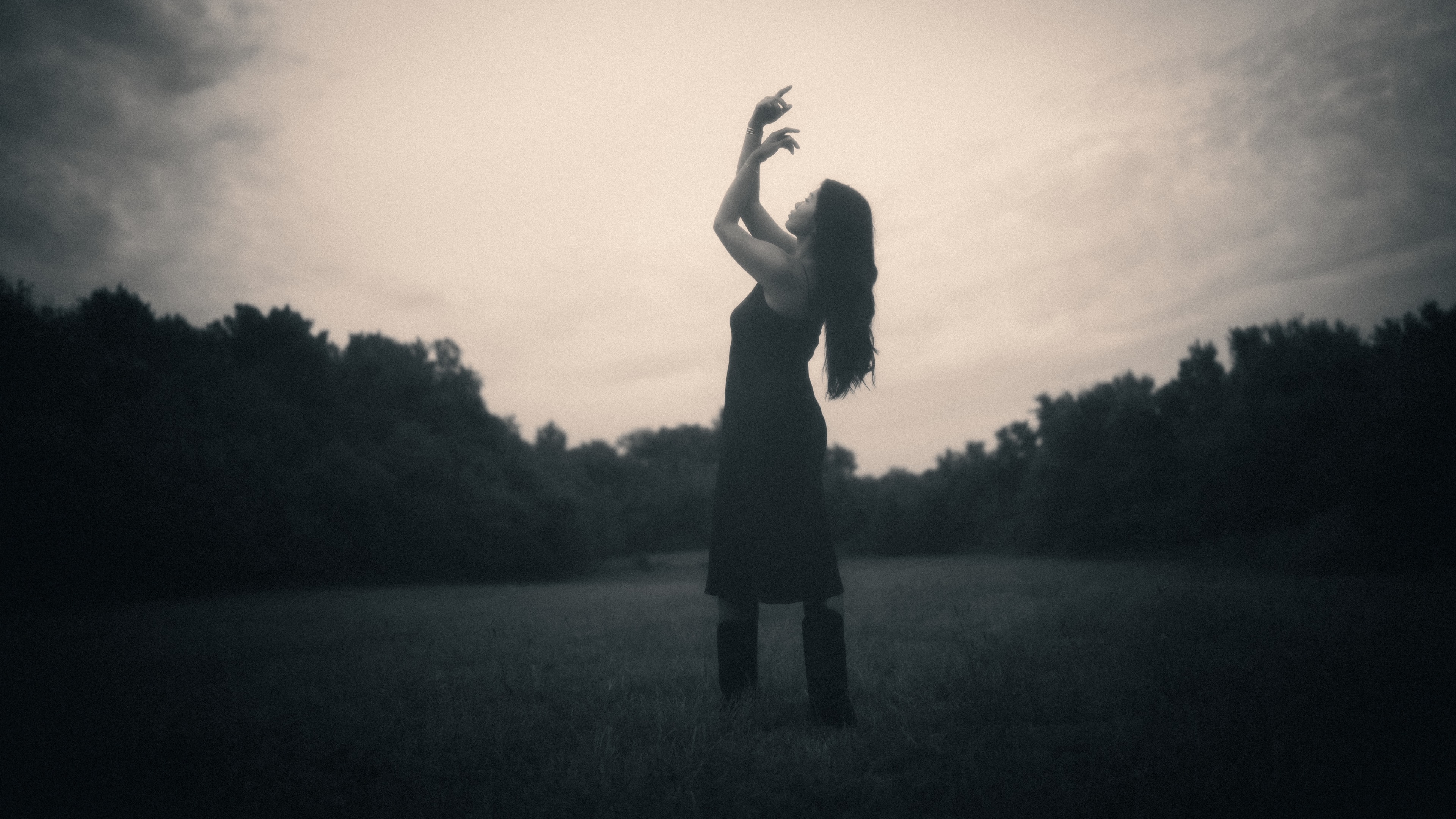 Silhouetted against a pale dusk sky, a long-haired woman in a sleeveless dress and knee-high boots tilts her head back with both arms raised, fingers curled skyward, framed by dark treelines in an open field.