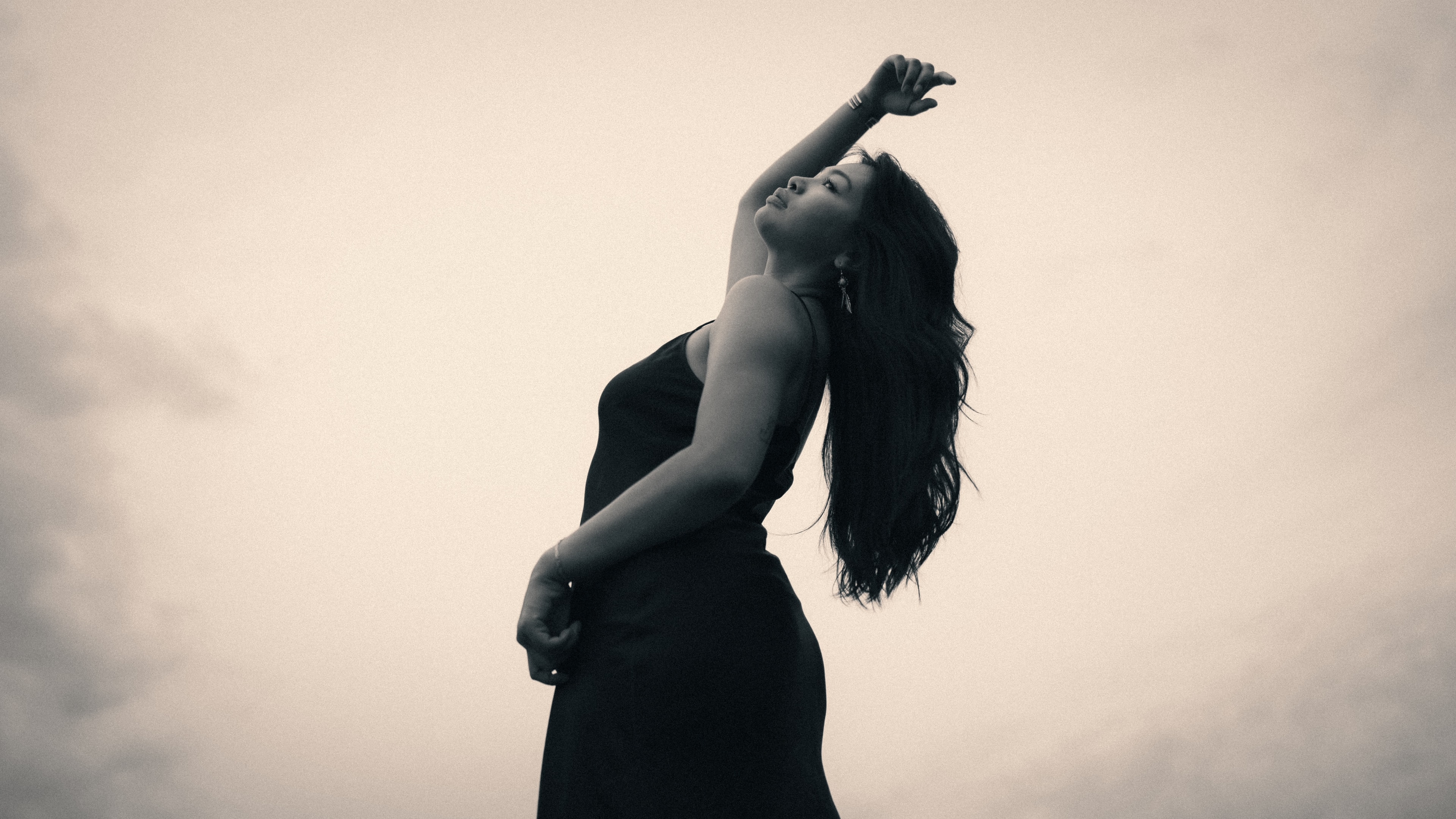 Side-profile silhouette of a woman in a black spaghetti-strap dress tilting her head back, one arm stretched skyward with hand curled, dark hair blown sideways across her face, low-angle shot against a hazy monochrome sky.