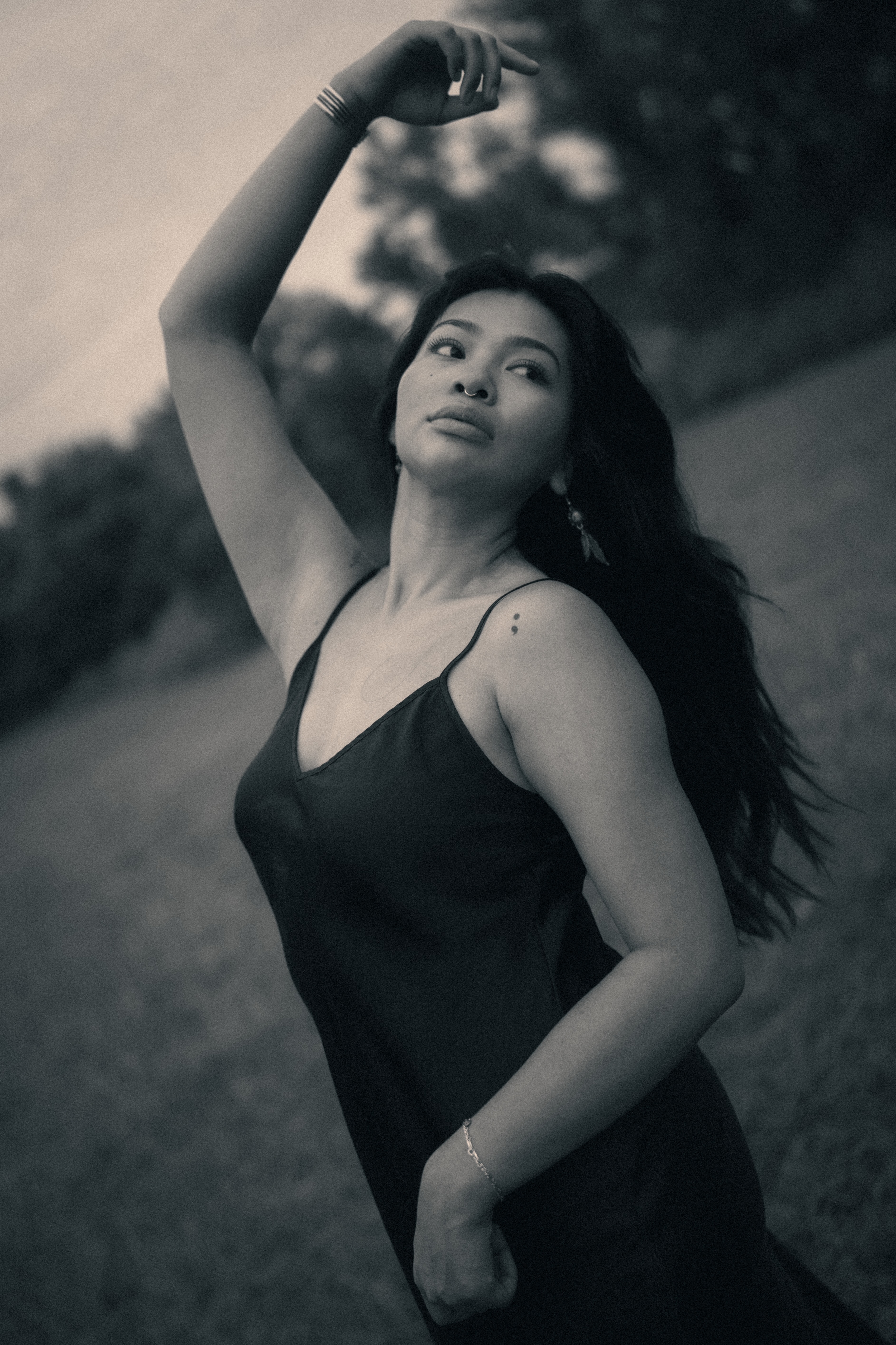 Black-and-white portrait of a woman with a septum ring and feather earring, one arm raised above her head, dark hair swept across her bare shoulder, wearing a black slip dress with delicate arm bracelets, standing outdoors against a blurred tree line.