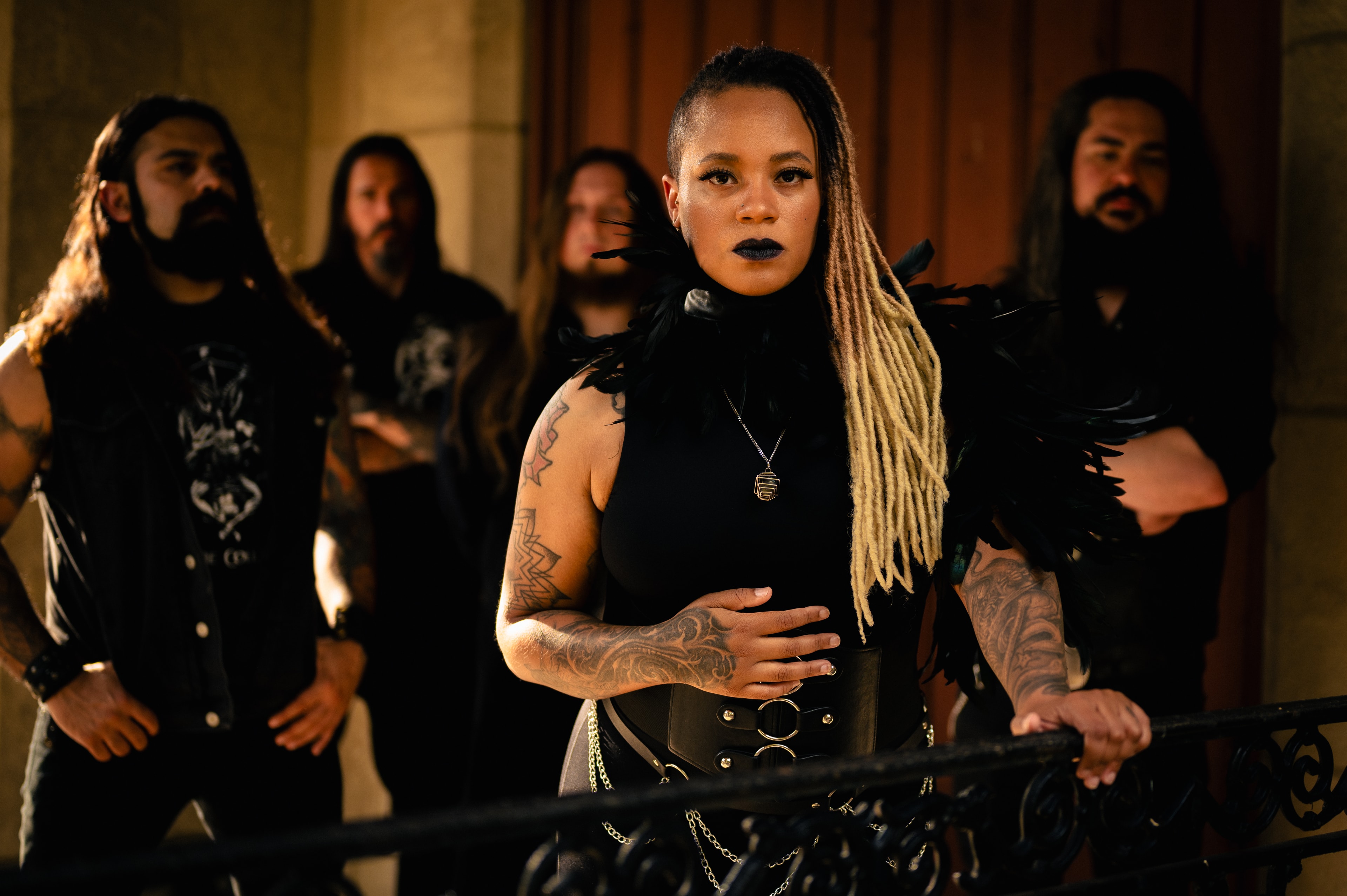 Female vocalist with black-to-blonde dreadlocks and dark lipstick rests one hand on her waist, wearing a black feathered collar and leather corset belt, four tattooed bandmates in black band tees blurred behind her on a stone building's steps at golden hour.