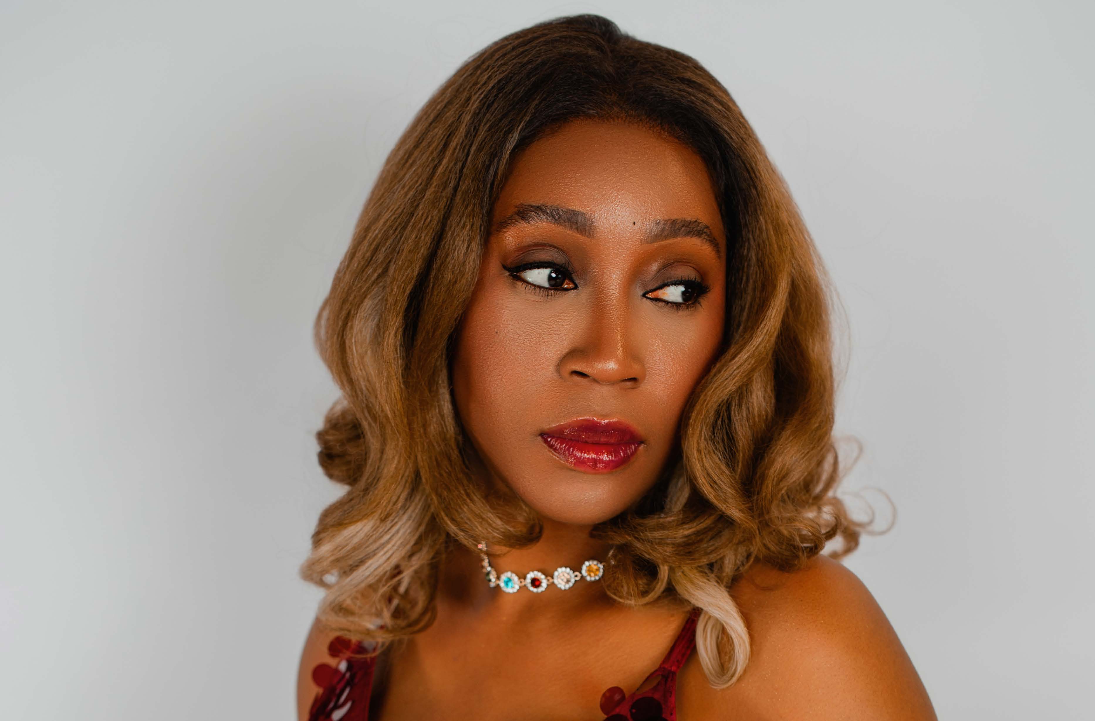 Close-up of a woman with voluminous curled bronde hair gazing to her left, smoky eyeliner and glossy red lips, wearing a beaded gemstone choker and off-shoulder red floral top against a plain gray backdrop.