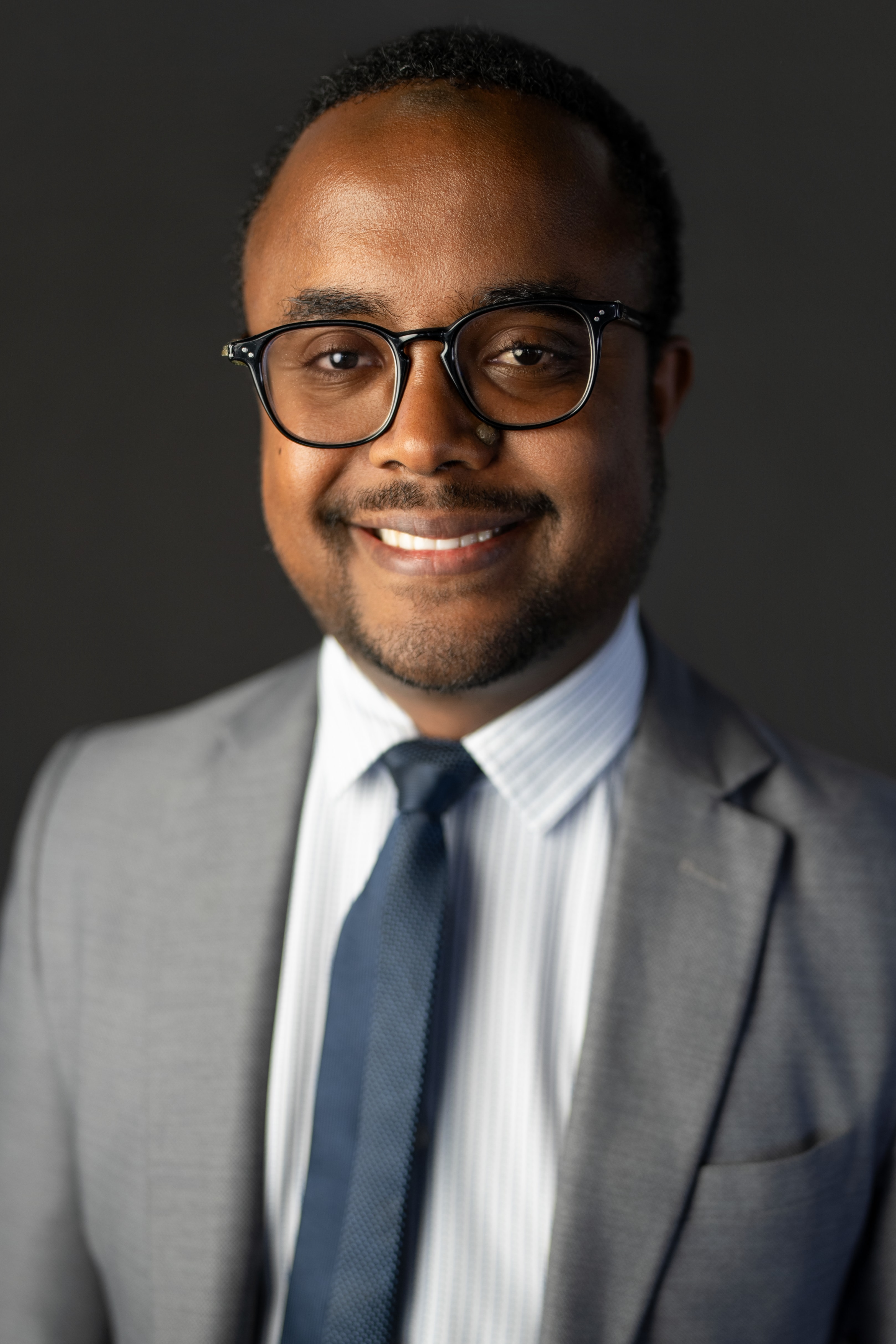 Close-cropped headshot of a mustached man in black-framed glasses and gray suit, dramatic side lighting illuminating his face against a nearly black background.