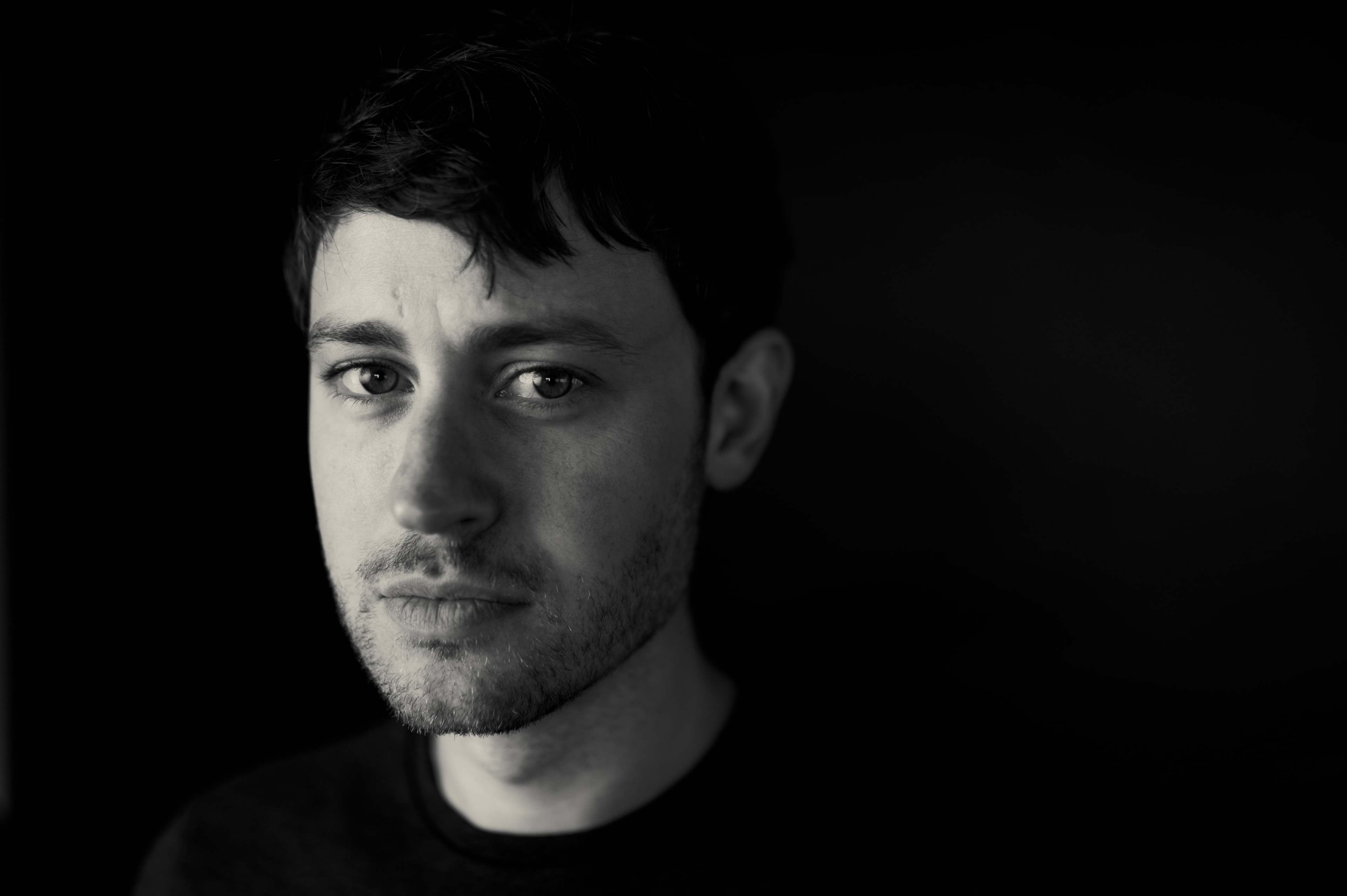 Black-and-white portrait of a man with dark tousled hair, faint forehead crease, and light stubble, dramatic side lighting on the left half of his face fading to black on the right, wearing a dark crewneck top against a pure black background.
