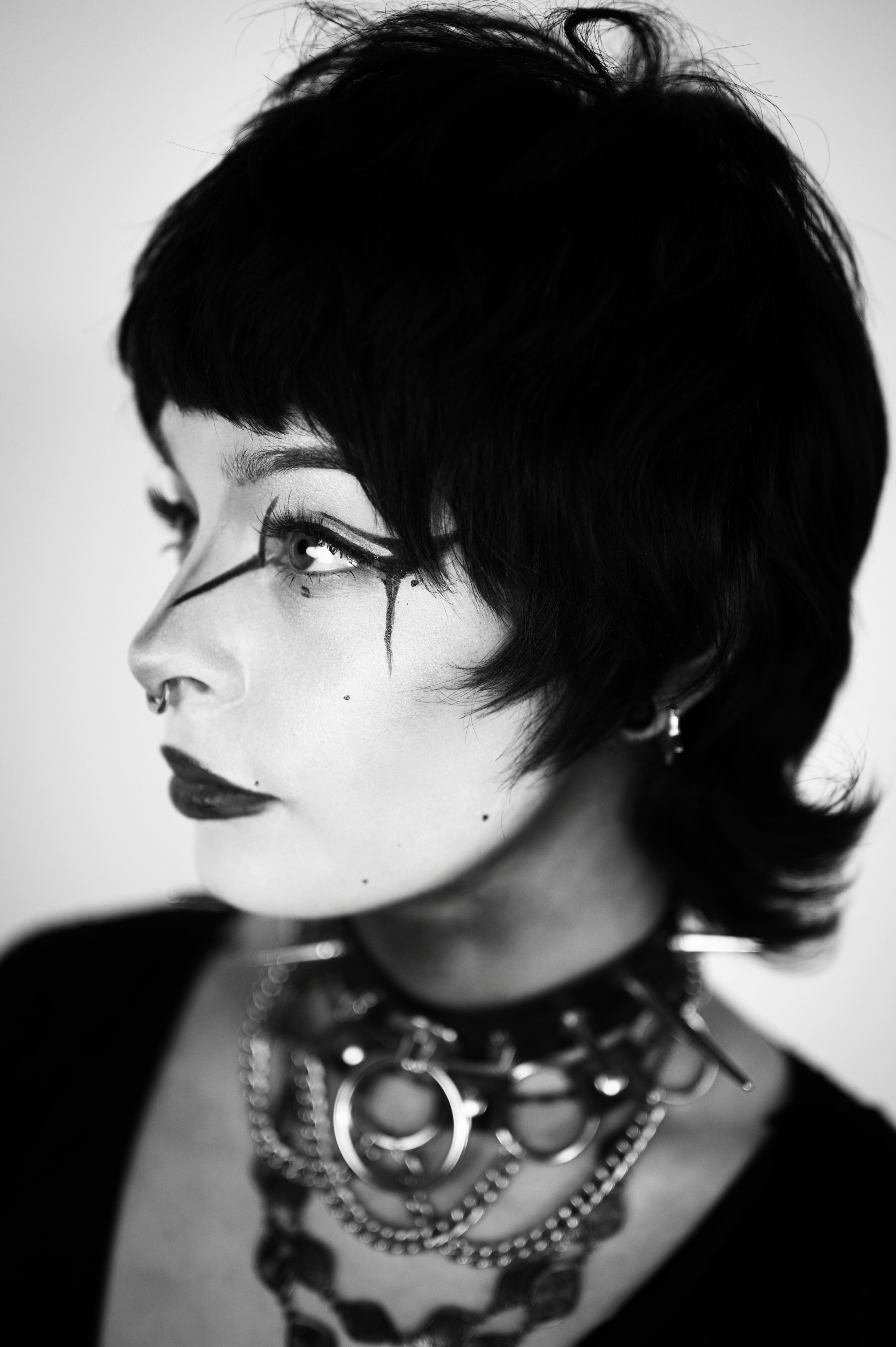 Black-and-white side-profile close-up of a woman gazing upward, sharp cat-eye liner with dripping lines under one eye, septum ring, dark lips, and a chunky chain-link choker layered with metal ring pendants against a soft white background.