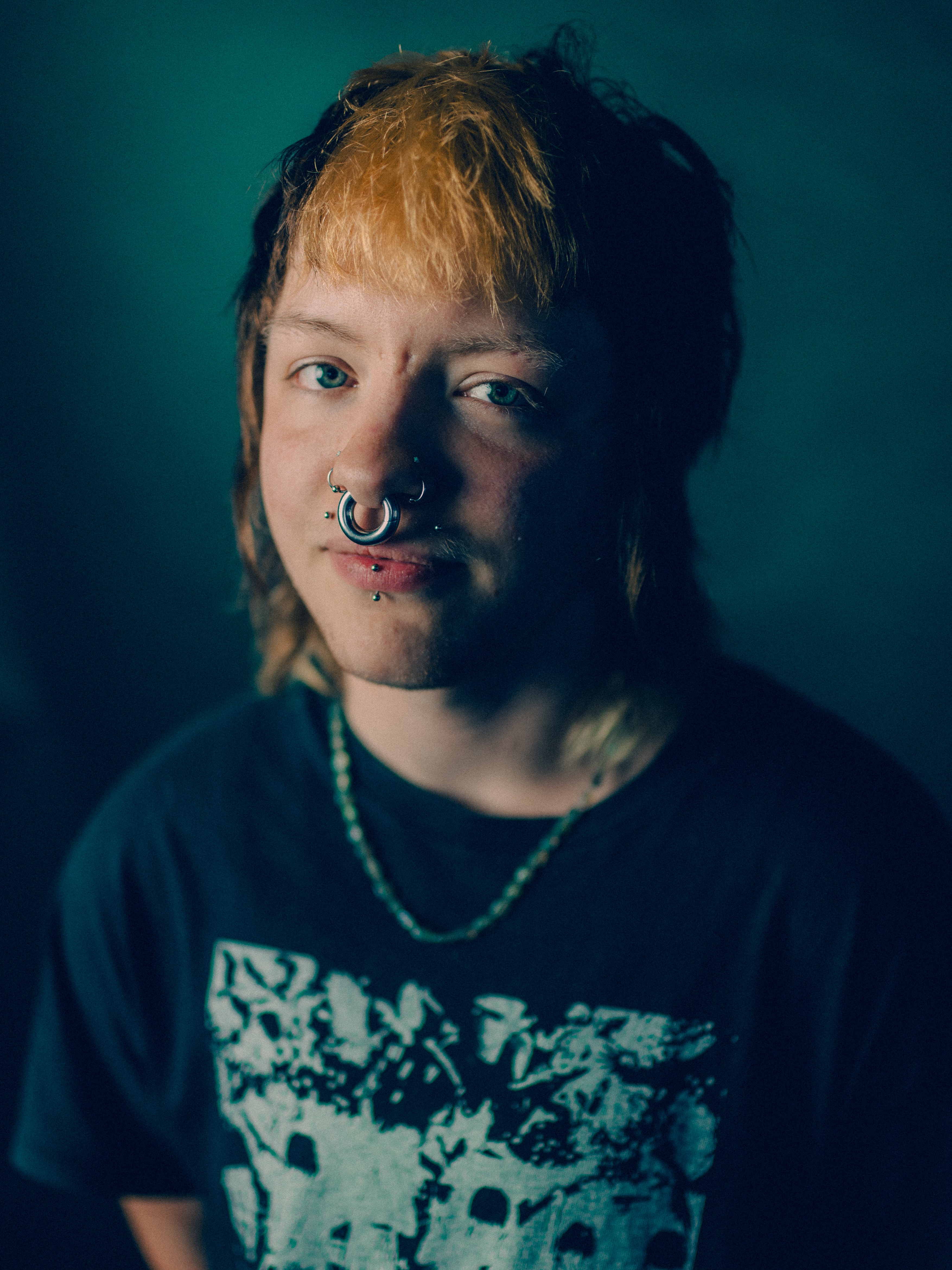Close-up portrait against a dark teal backdrop of a young man with a two-tone mullet, orange-bleached fringe over dark roots, septum ring, chin and cheek piercings, and a faint scar above his eyebrow, dramatic side lighting casting half his face in shadow, wearing a black graphic tee and a beaded chain necklace.