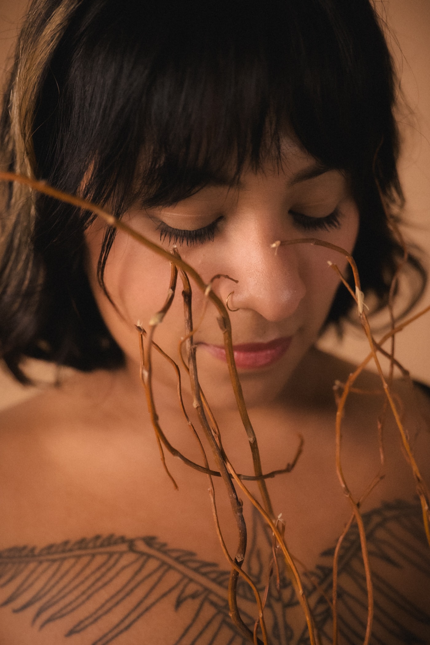 Close-up of a woman with dark blunt-cut bangs, eyes closed and chin tilted down, nose ring visible, thin dried vines hanging vertically across her face and fern-tattooed shoulder, warm amber lighting.