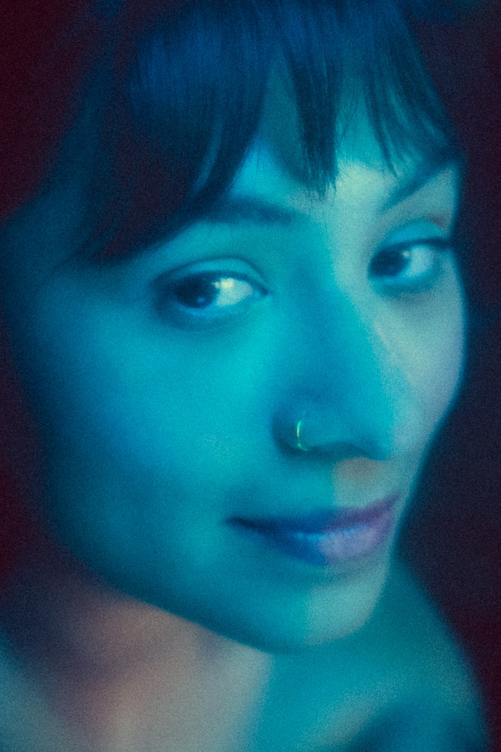 Extreme close-up of a woman's face lit in split teal-blue tones, dark eyes glancing down-left, faint smile, small hoop nose ring catching the light.
