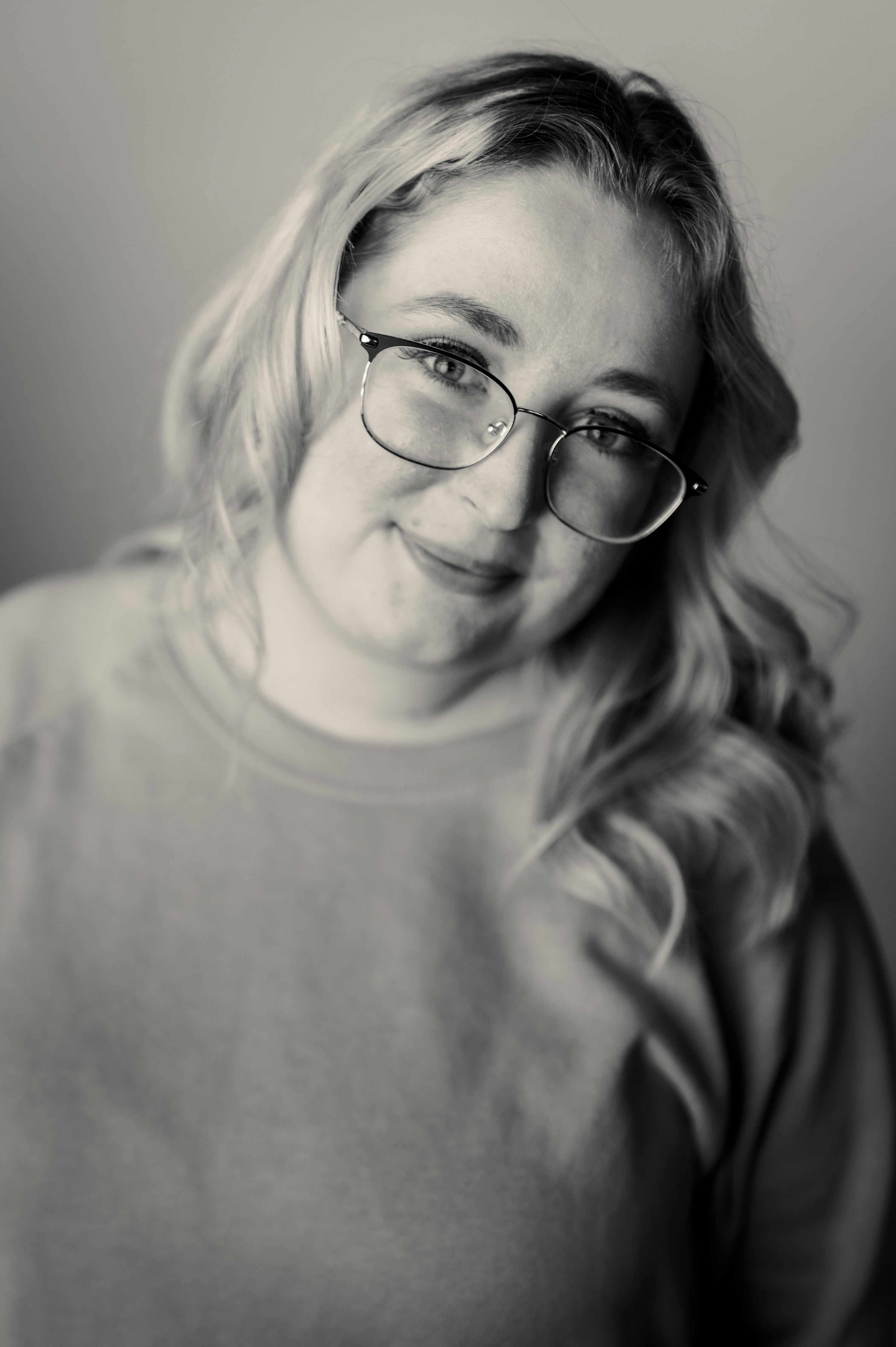 Black-and-white portrait of a woman in wire-frame glasses and an oversized sweater, head tilted sharply right, wavy hair cascading past one shoulder, soft window light on her face against a plain backdrop.