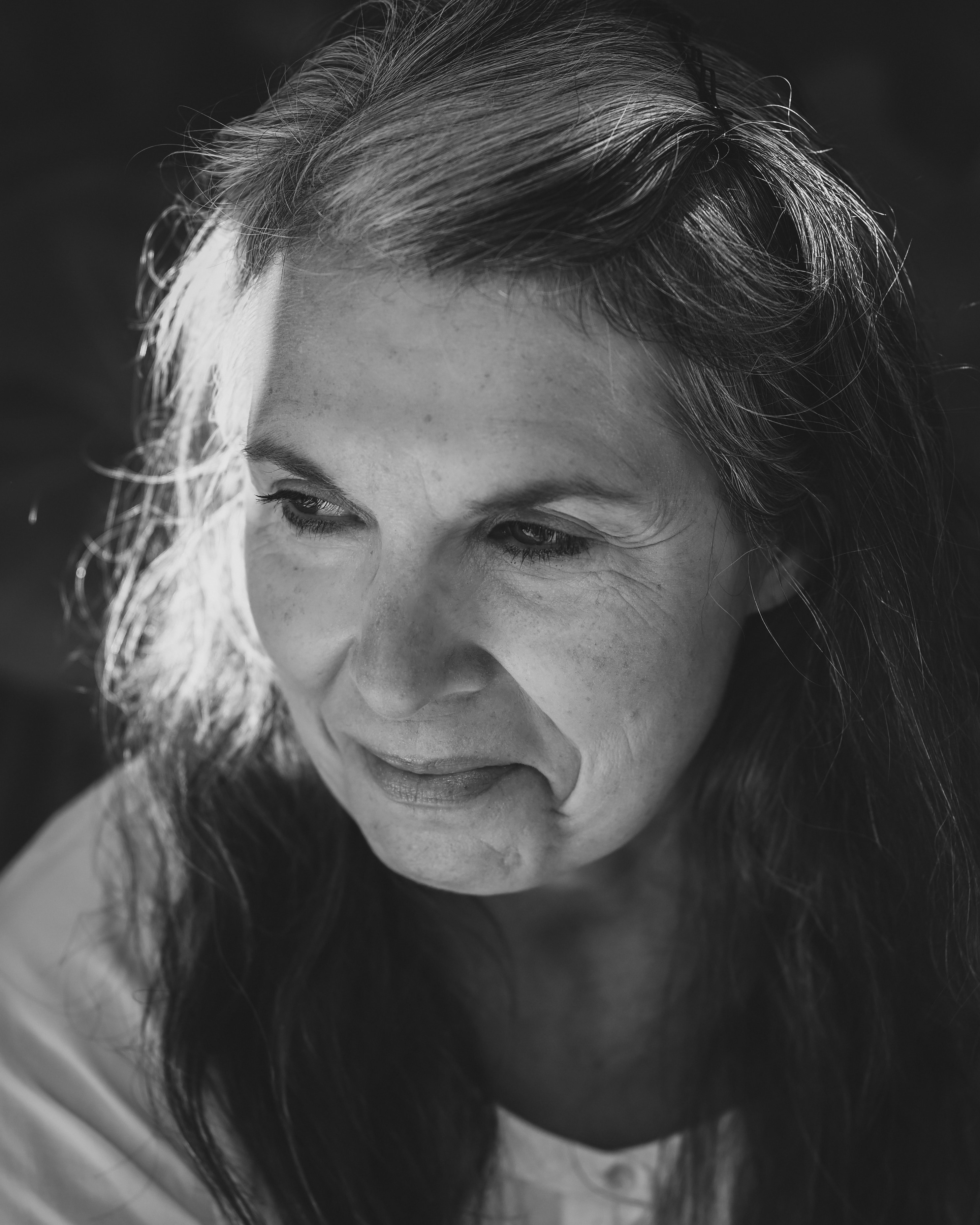 Black-and-white close-up of a woman with long wavy dark hair, eyes cast down and slight smirk, sunlight rimming one side of her hair against a black background, wearing a white top.