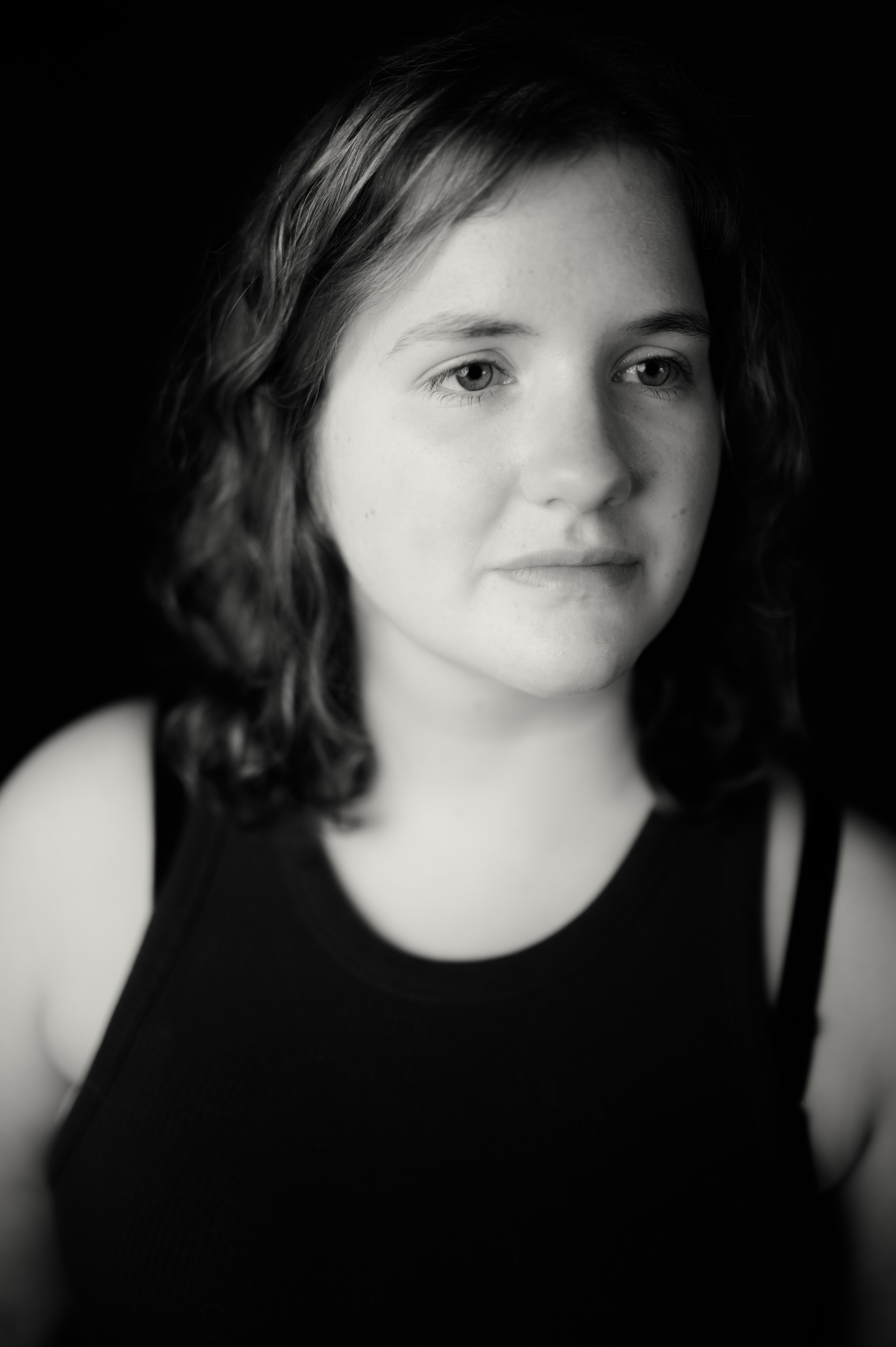 Black-and-white close-up of a young woman with wavy hair parted to one side, gazing off-camera with a subtle closed-mouth expression, dramatic side lighting fading into a black background.