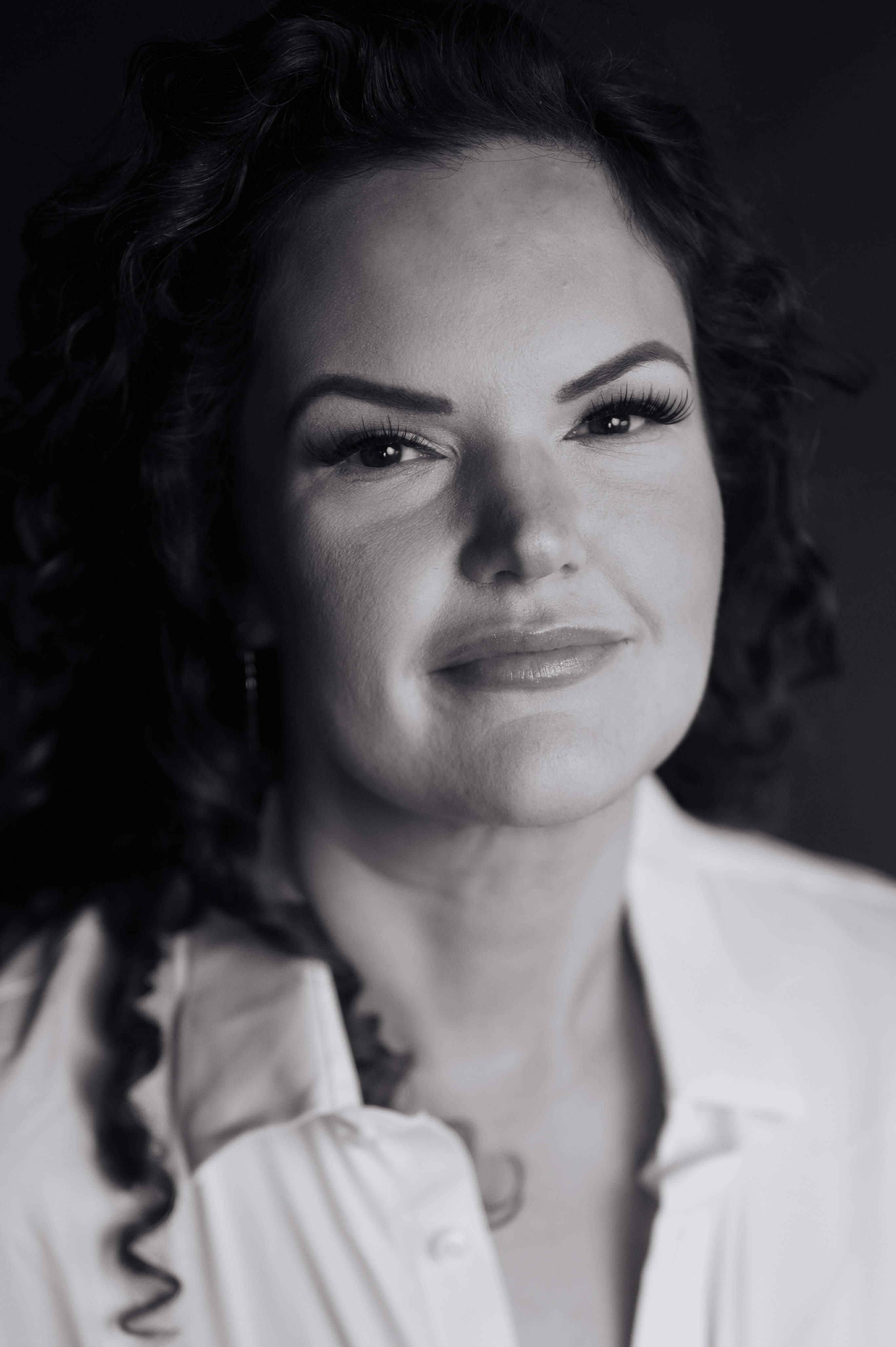 Black-and-white close-up portrait of a woman with voluminous curly dark hair, chin tilted up, faint smile, wearing an open white collared shirt against a dark background with strong side lighting sculpting her cheekbone.