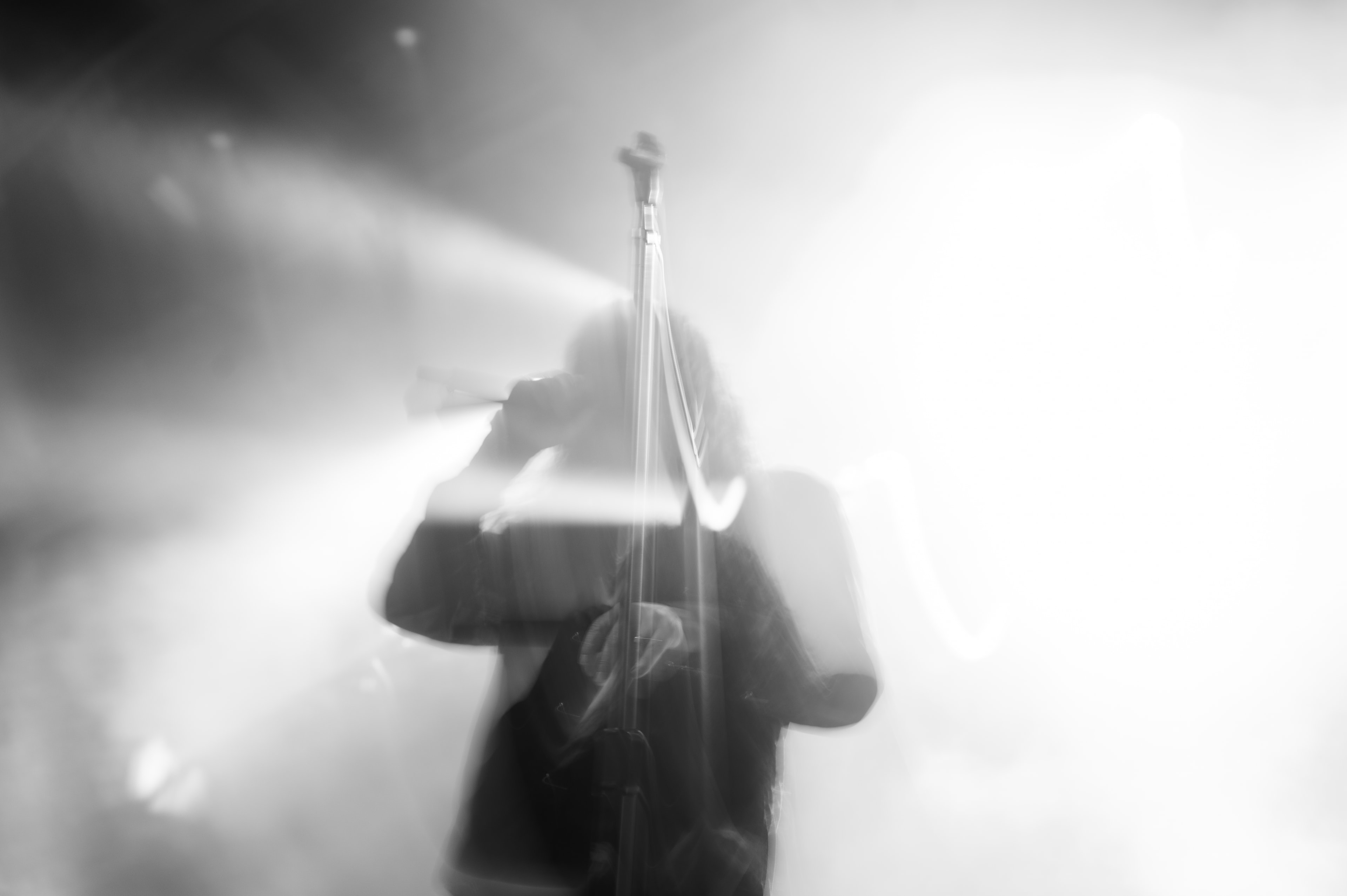 Long-exposure black-and-white stage shot of a singer's blurred silhouette leaning into a mic stand, streaked light trails crossing the frame amid dense white haze.