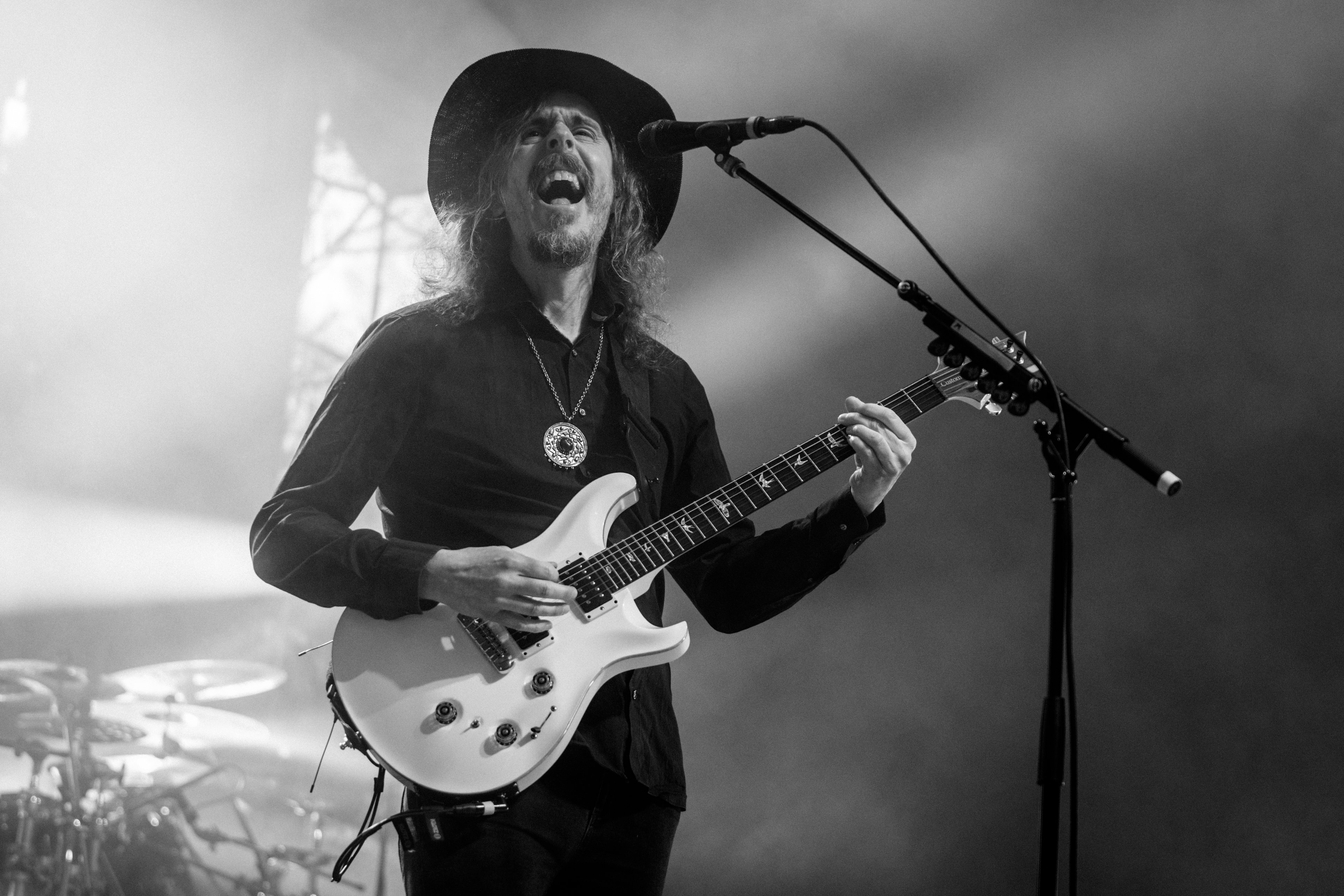 Long-haired musician in a black wide-brimmed hat, mouth open mid-shout, gripping a white PRS guitar neck beside an angled boom microphone in hazy monochrome stage light.