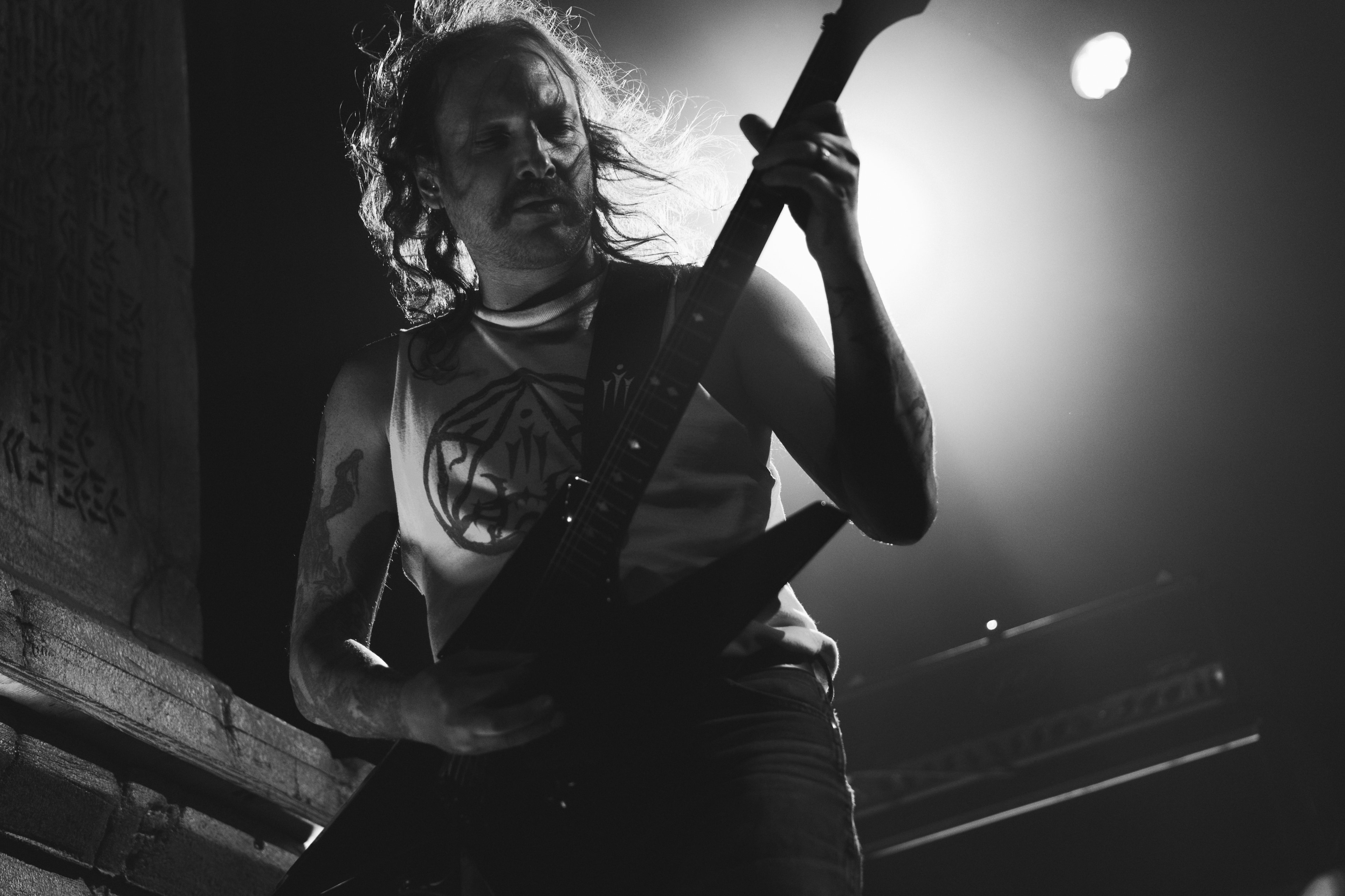 Low-angle black-and-white shot of a shaggy-haired guitarist gripping a dark flying-V guitar, hair blown mid-motion, silhouetted against a bright circular stage light.