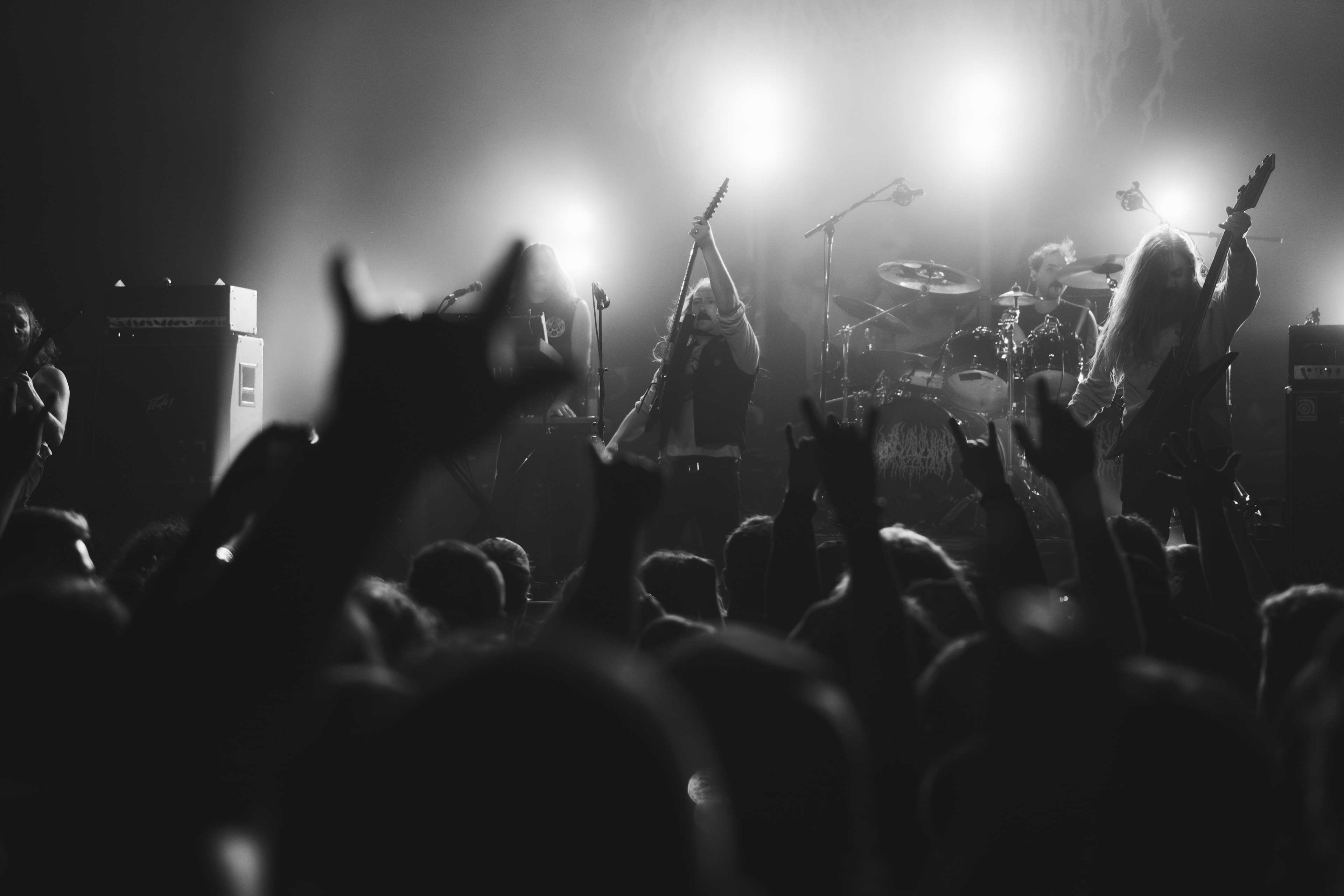 Black-and-white stage shot from behind the crowd: silhouetted hands raised in horns, a guitarist lifting his instrument overhead, long-haired bassist and drummer lit by three hazy backlights.