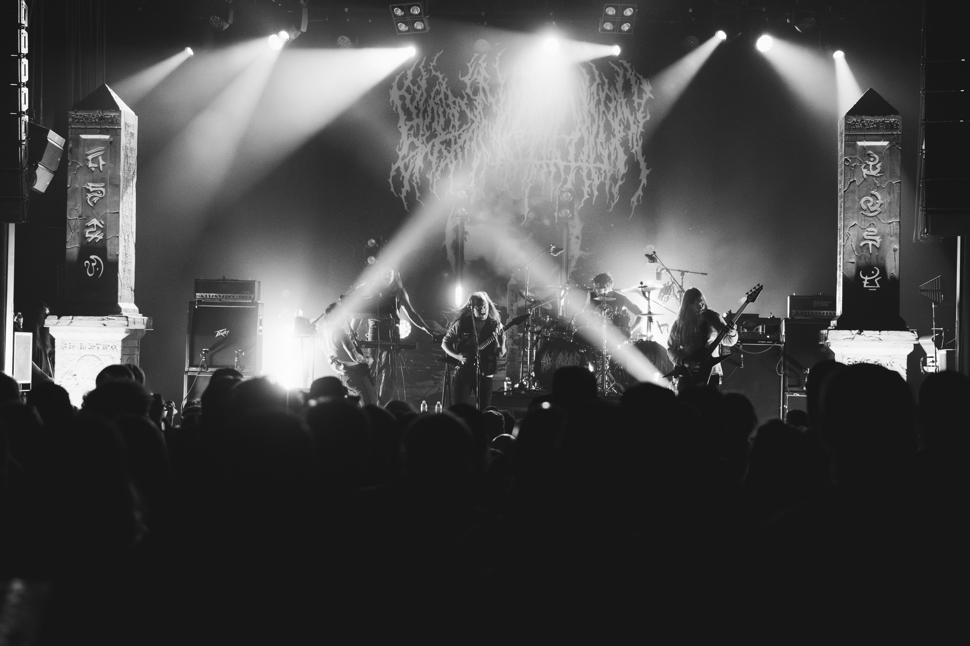 Black-and-white stage shot of four musicians spread across a wide stage flanked by two tall carved obelisk pillars, guitarist and vocalist at center mics, drummer backlit behind cymbals, crisscrossing light beams cutting through haze above a silhouetted crowd of raised phones in the foreground.