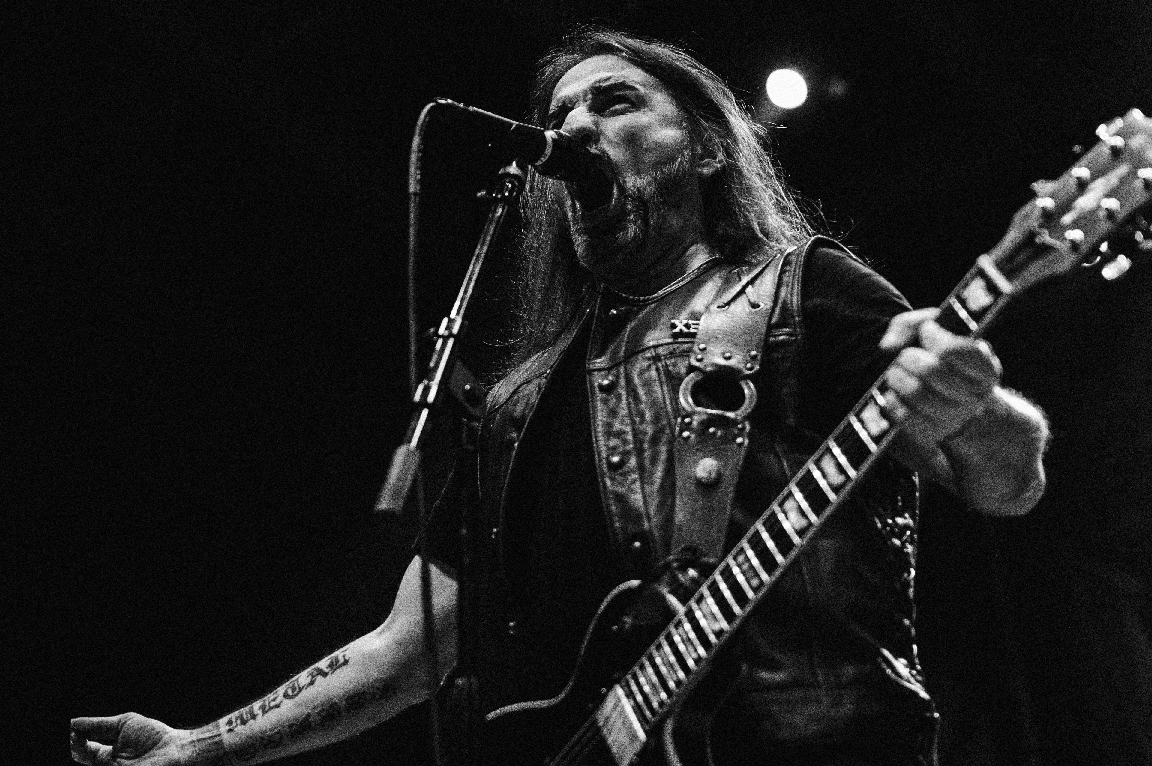 Black-and-white low-angle shot of a bearded, long-haired musician screaming into a mic stand while gripping an angled guitar neck, "metal" tattooed on his raised forearm, a single round stage light glowing over his shoulder.