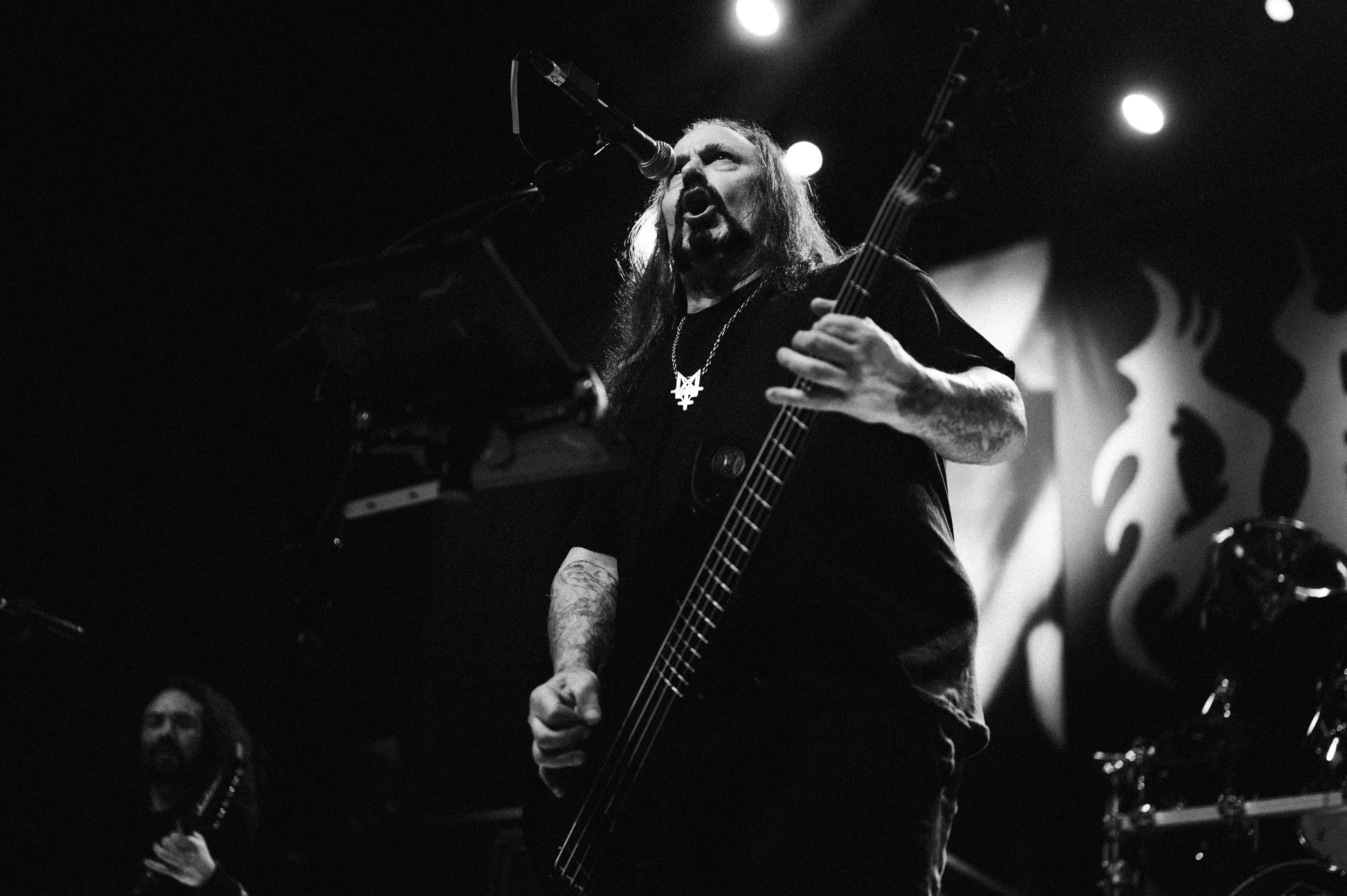 Bearded, long-haired bassist mid-scream at a low-angle stage shot, tattooed forearm gripping the fretboard, drum kit and flame-patterned backdrop blurred behind him.