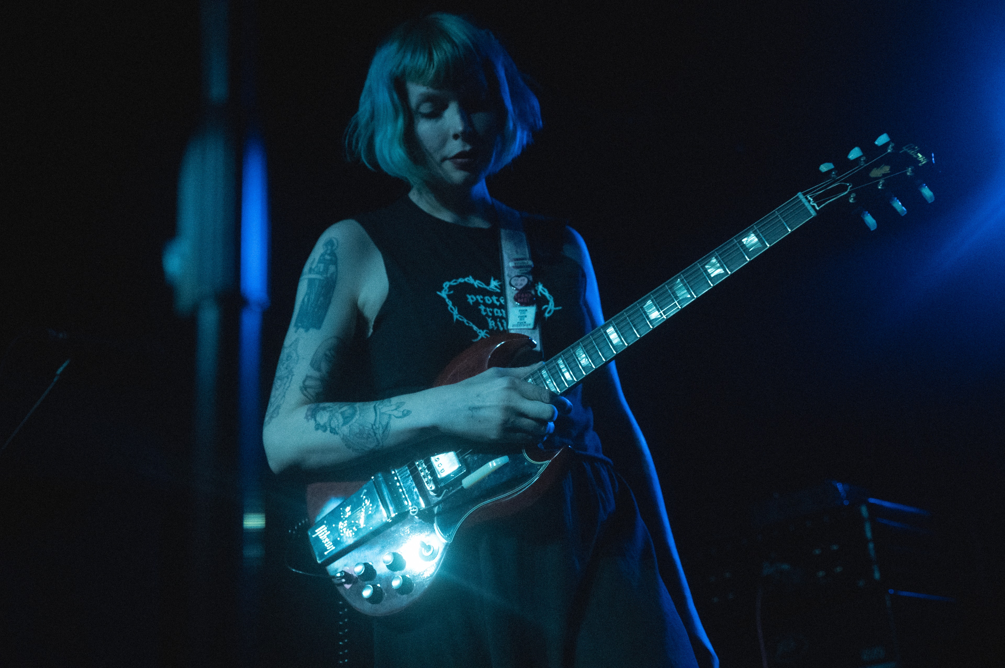 Bob-haired guitarist in a black graphic tank top plays a red guitar with an illuminated effects unit clipped to the strap, bathed in blue stage light, eyes cast down toward the fretboard.