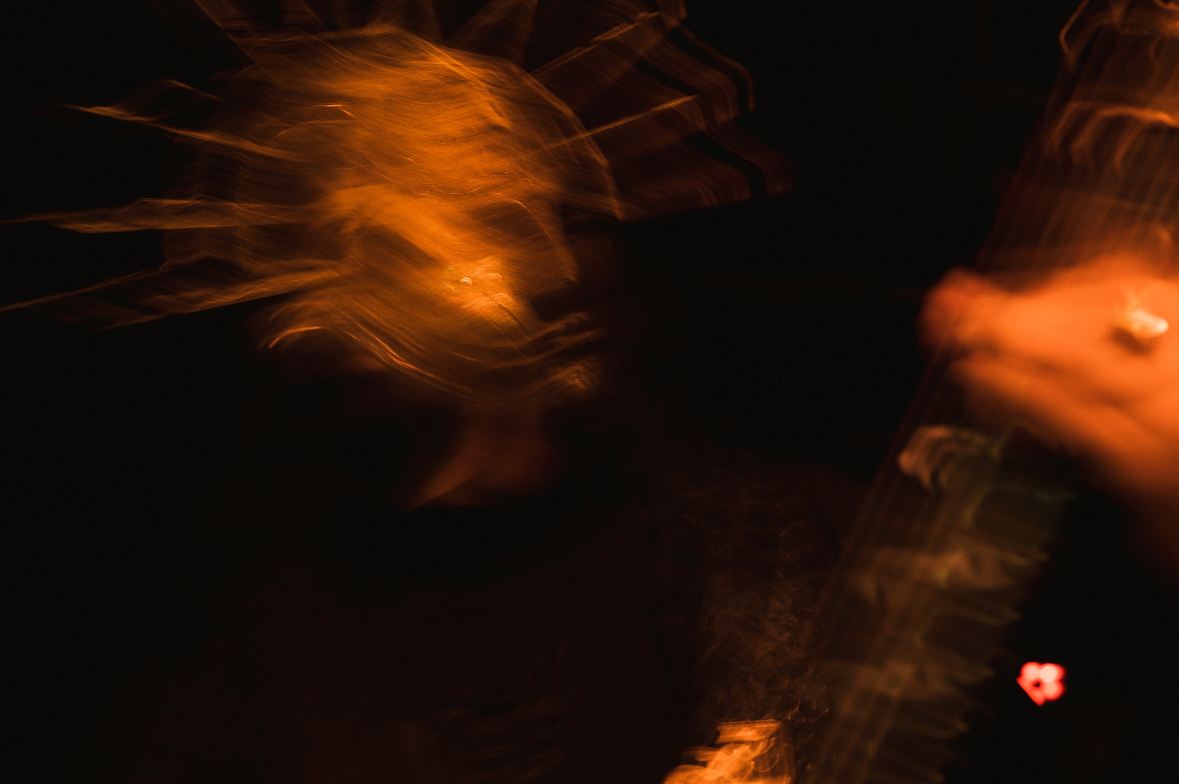 Long-exposure blur of a musician swinging their head, orange stage light trailing through hair like flames, a hand gripping a guitar neck in the dark foreground, small red light glowing bottom right.