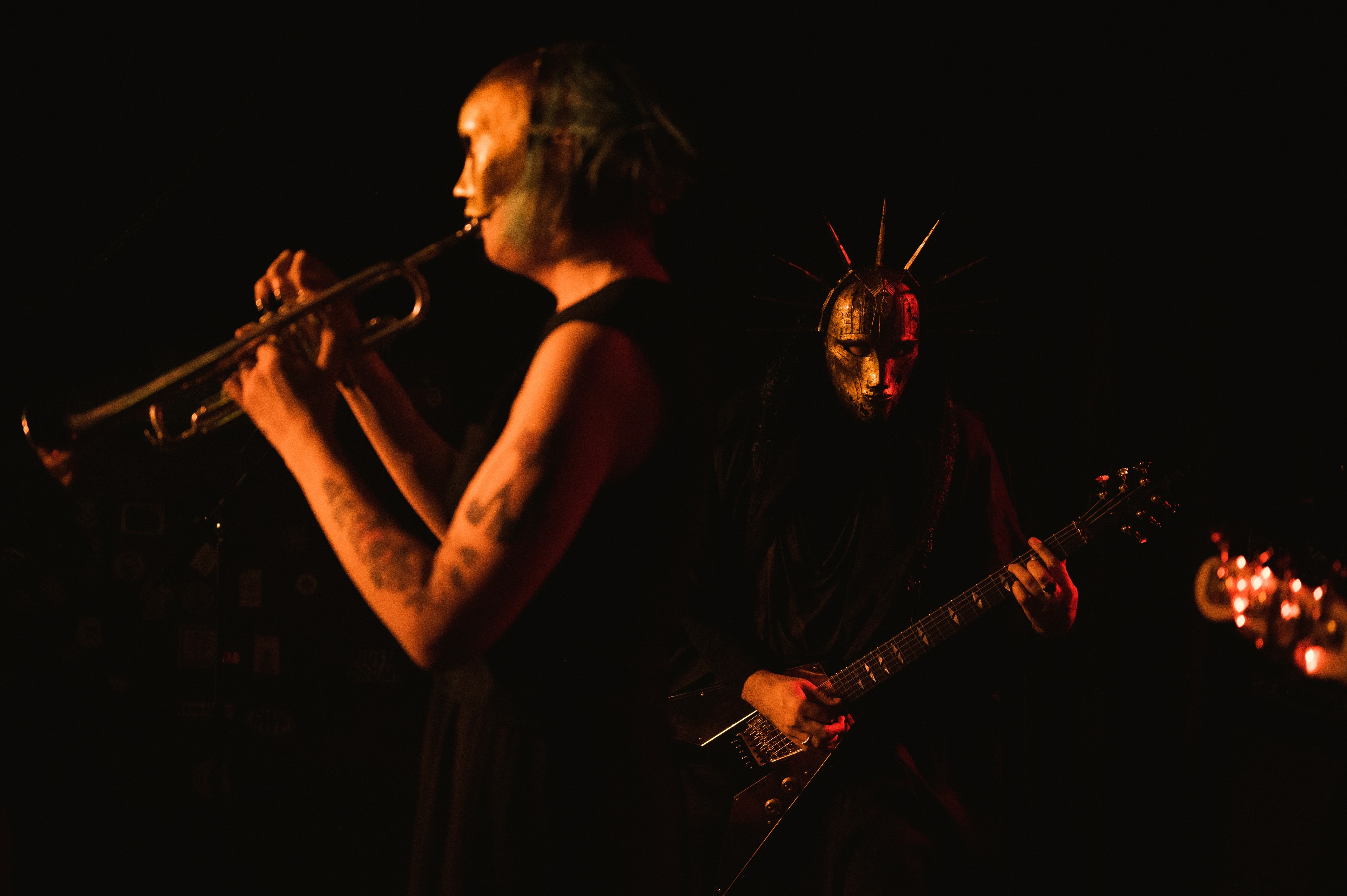 Trumpet player with green-painted face and shaved head plays mid-note beside a spike-crowned, mask-wearing guitarist lit in dim red-orange stage light.