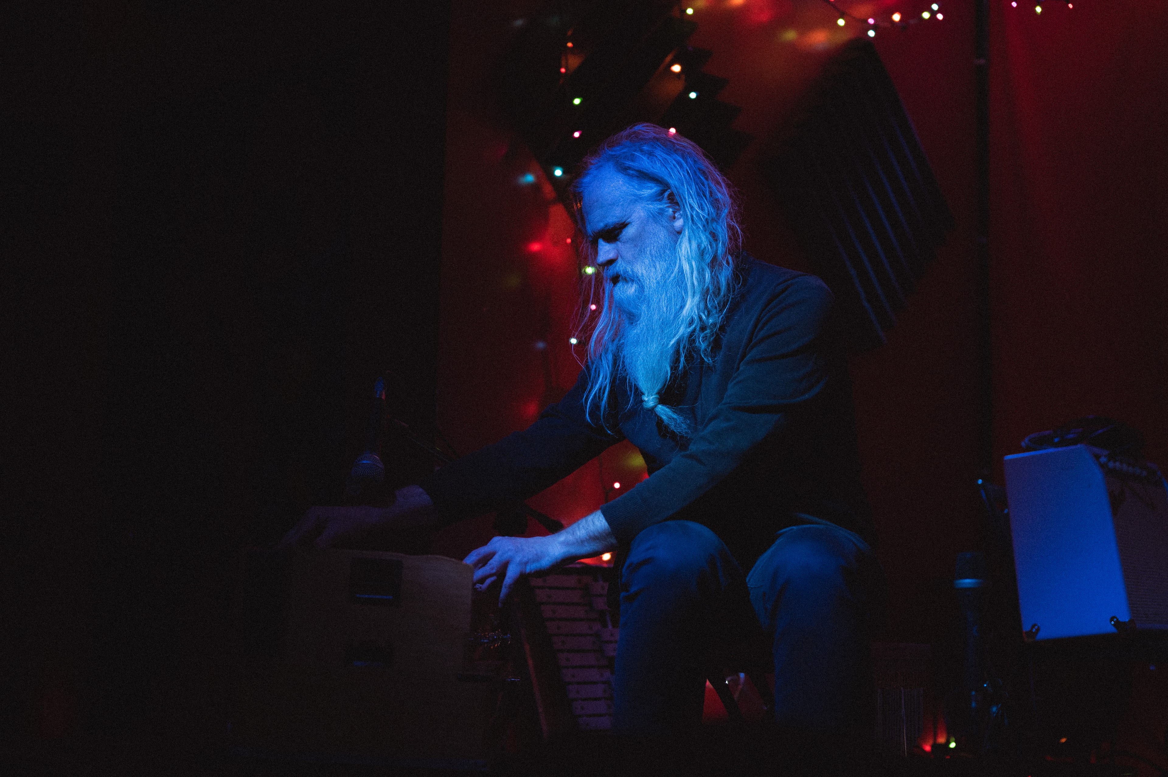 Bearded man with long gray hair bent over a wooden cajon-like instrument on stage, blue lighting casting his profile against a red wall strung with colorful string lights.