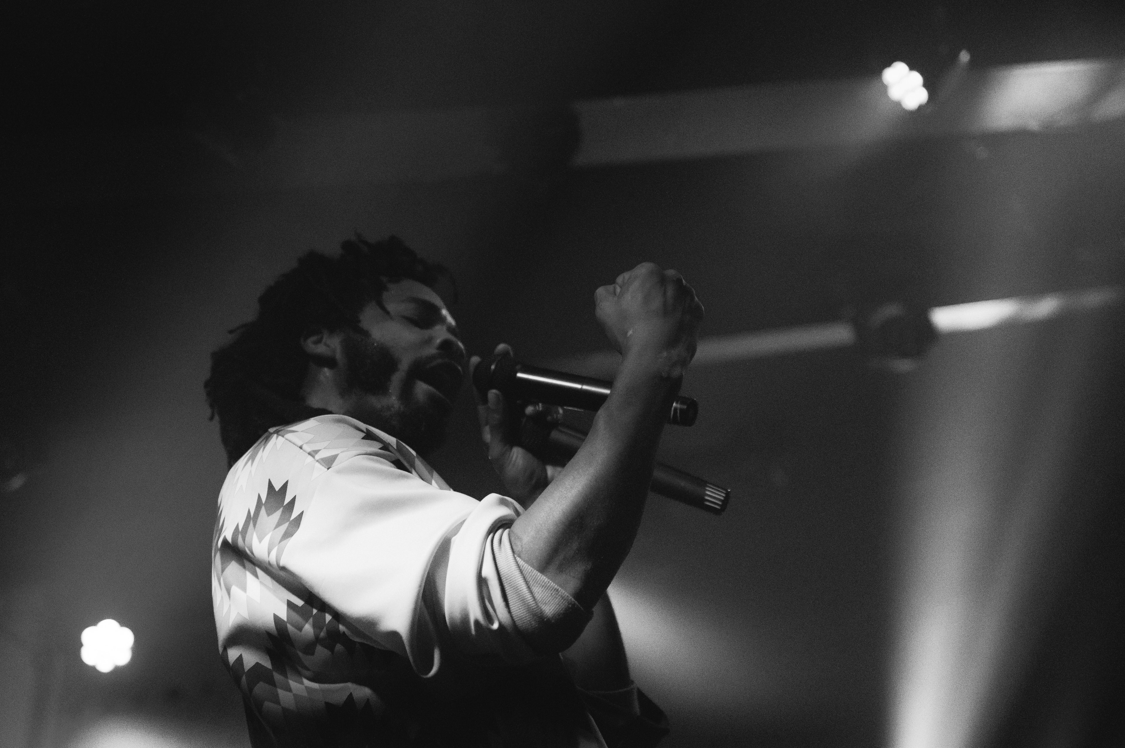 Low-angle shot of a bearded singer in a geometric-print jacket, eyes closed, gripping a mic with raised fist as stage lights streak diagonally overhead.
