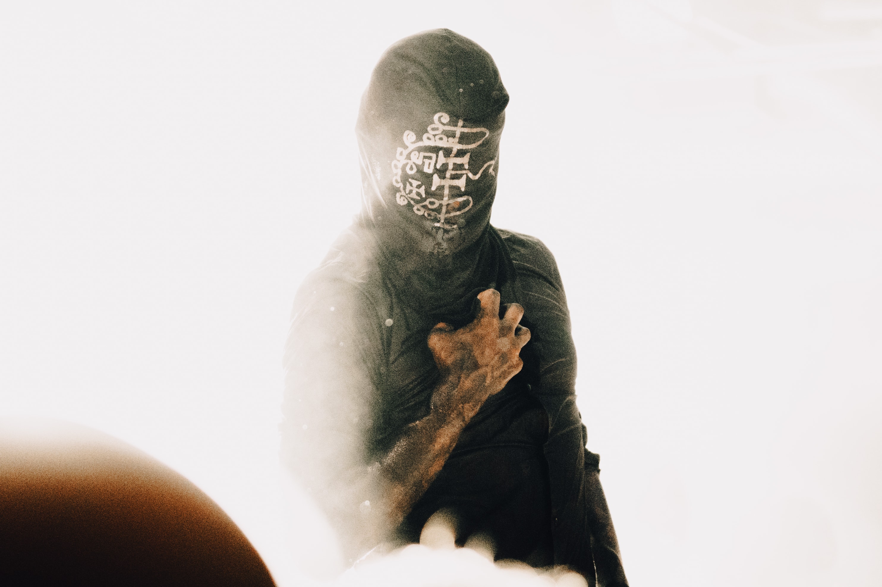 Person in a dark hood and mask painted with white sigil-like symbols across the face, gripping their chest with a soiled hand, backlit by bright white haze with a dark blurred orb in the foreground.