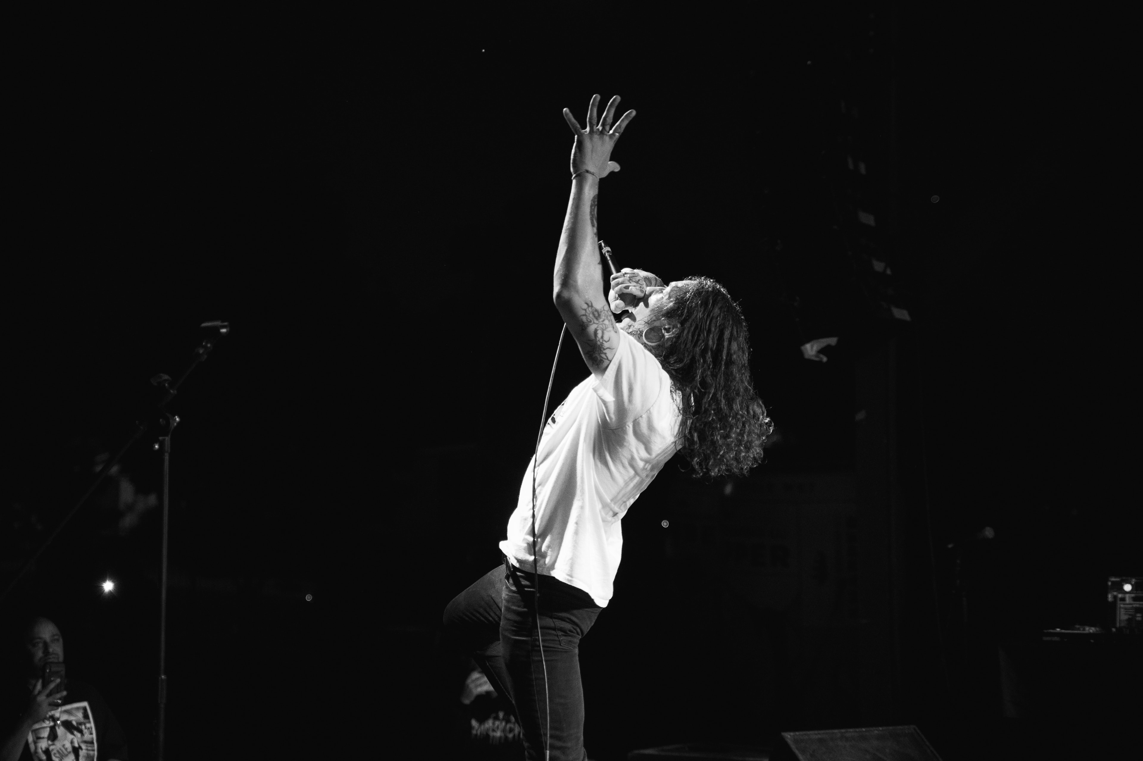 Black-and-white stage shot of a shirtless-armed singer in a white t-shirt and dark jeans, bent backward with one tattooed arm raised and fingers splayed, mic cable trailing down, silhouetted figures faintly visible in the dark foreground.