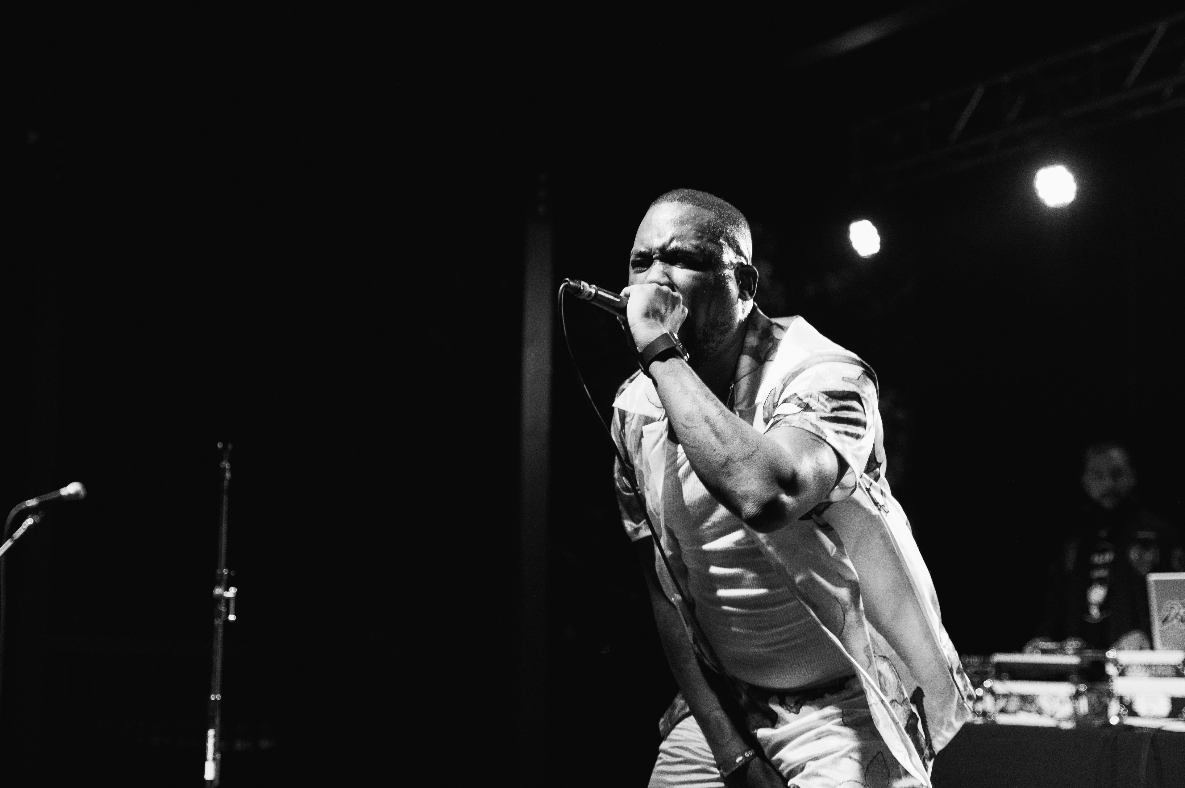 Bald male performer clenching a corded microphone to his mouth with a wristband-clad fist, brow furrowed, satin floral jacket open over a tank top, spotlight cutting through darkness behind him.