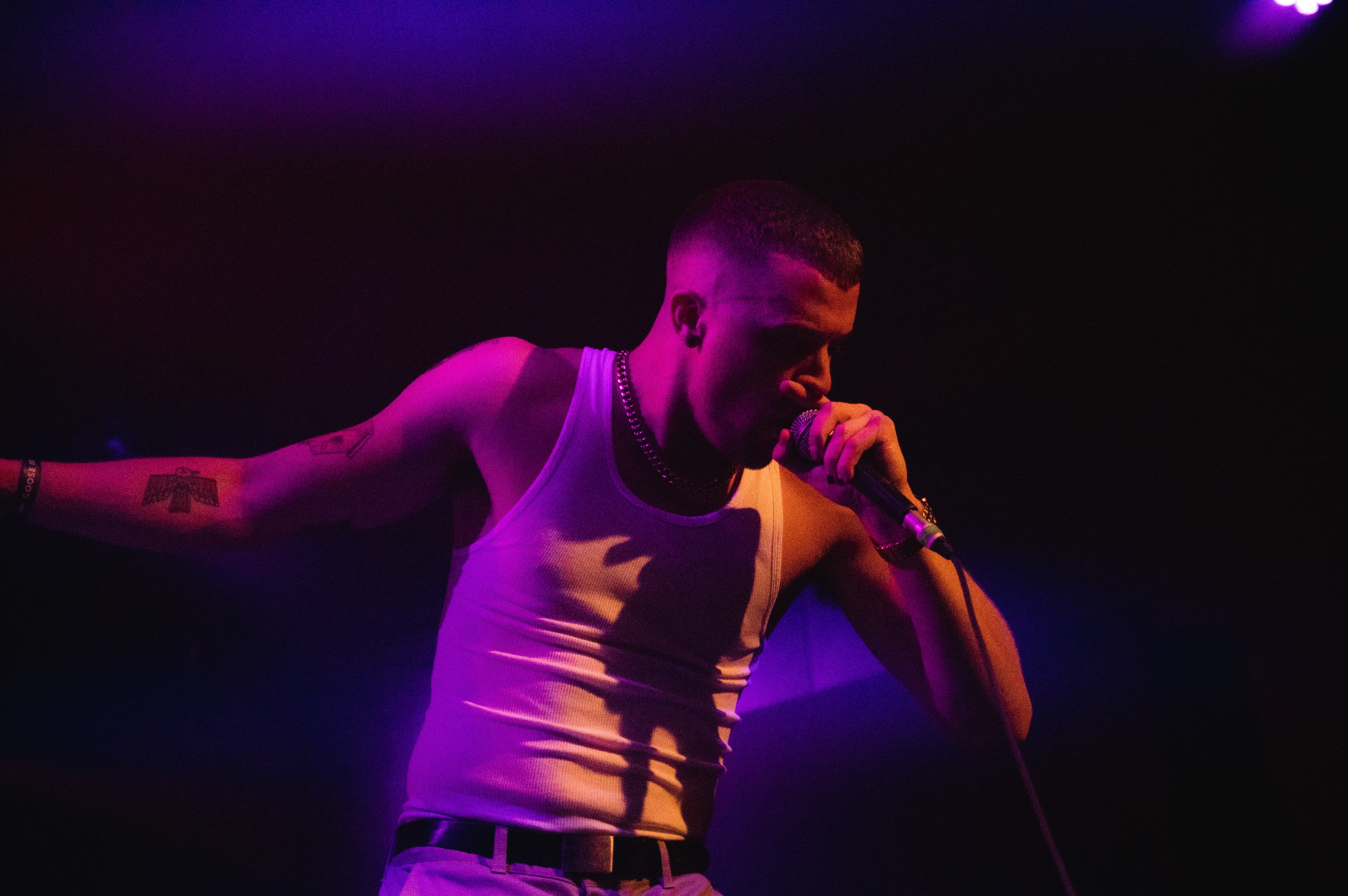 Male performer with a bird-and-rattlesnake tattoo on his forearm sings into a microphone, head tilted down, one arm outstretched, under vivid purple stage lighting with a chain necklace visible.