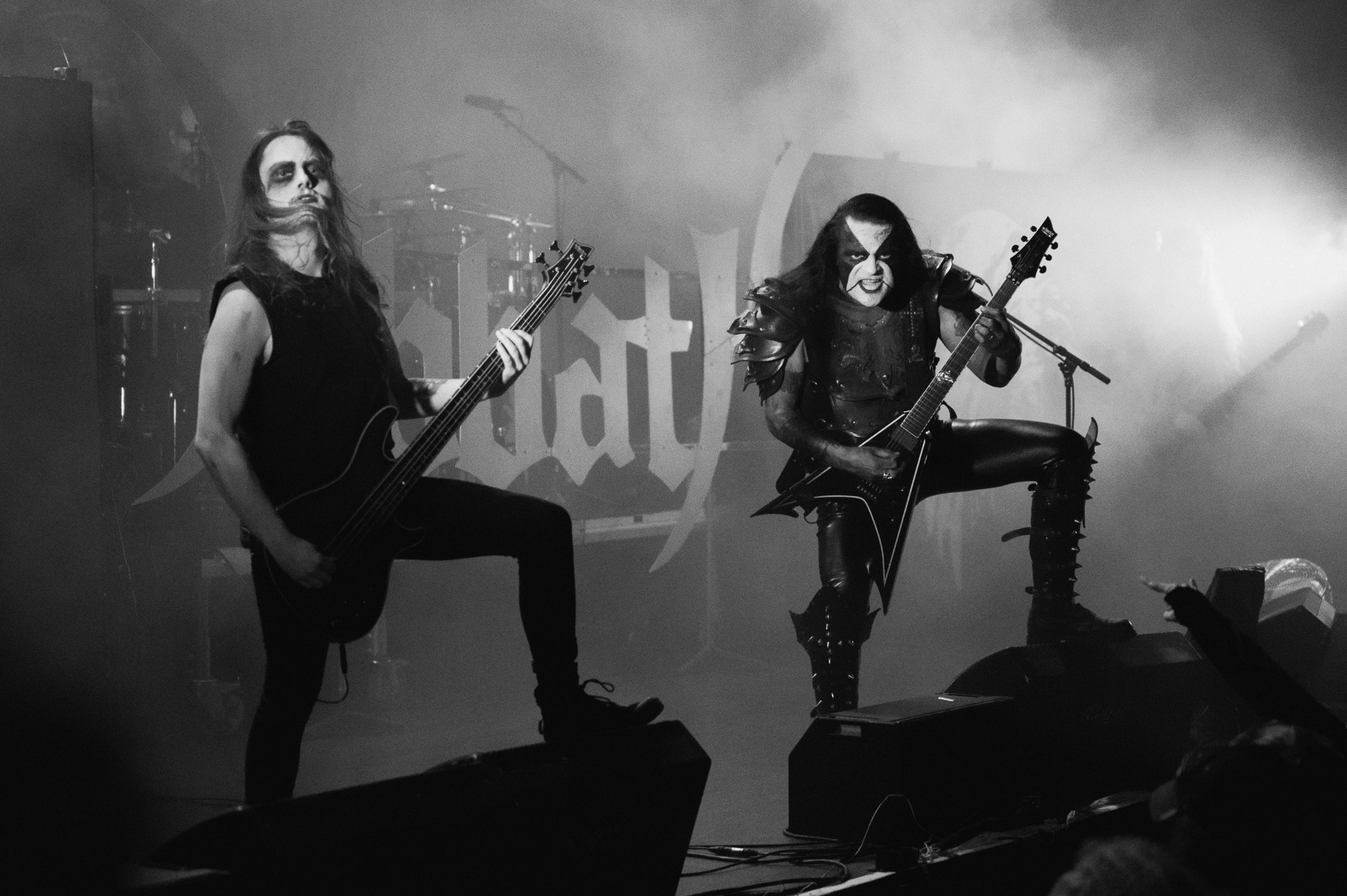 Two corpse-painted black metal musicians onstage, one in spiked shoulder armor and tall spiked boots striking a wide-legged pose with a V-shaped guitar, the other in a sleeveless black shirt playing bass with foot propped on a monitor, thick fog and backlit haze behind a partly obscured band logo.