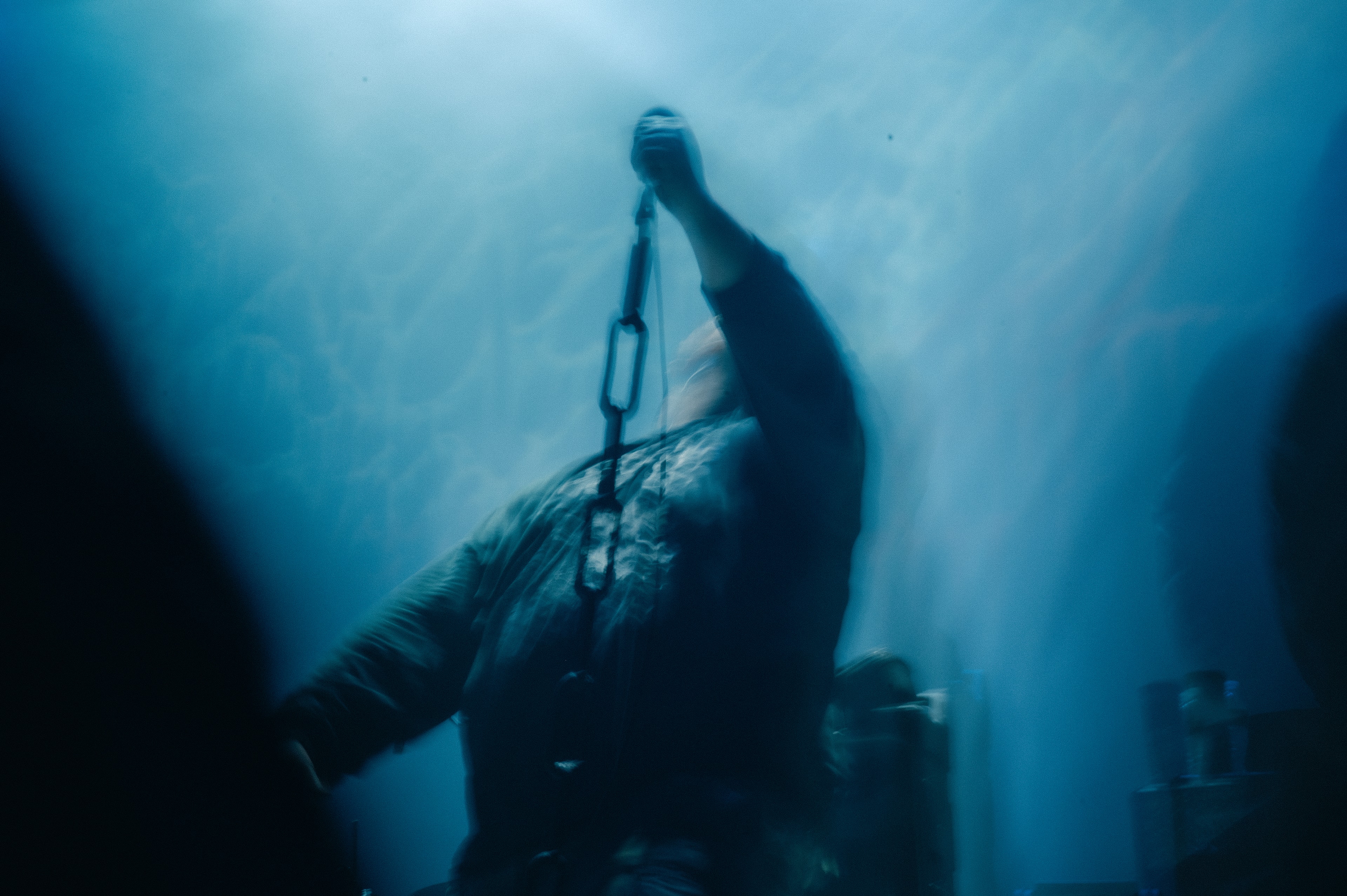 Silhouetted singer on stage tilting head back, arm raised high gripping a chain-suspended microphone, blurred with streaks of blue haze light behind.