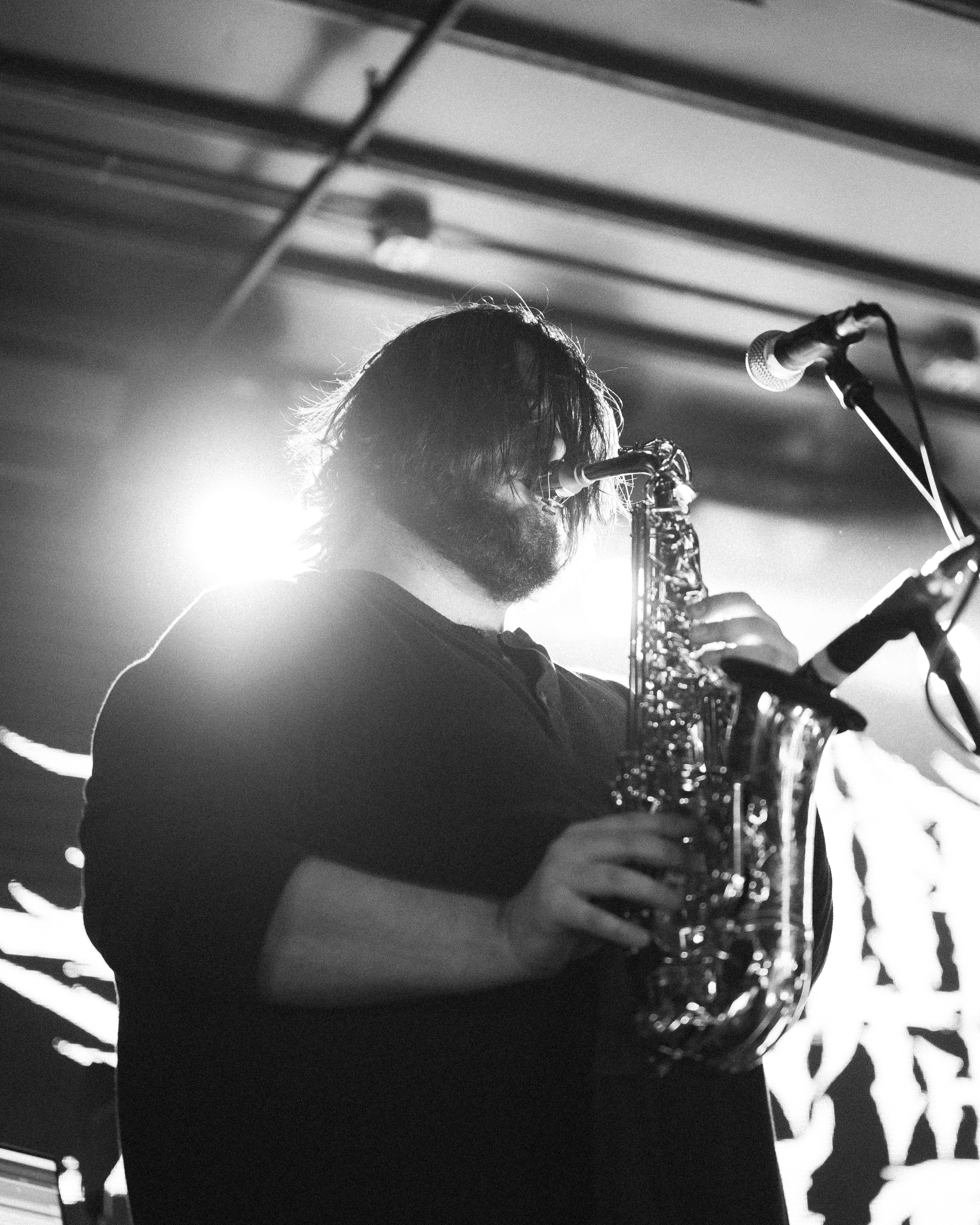 Bearded saxophonist with shaggy hair bowed over his instrument, silhouetted against a bright backlight beside two stage microphones.