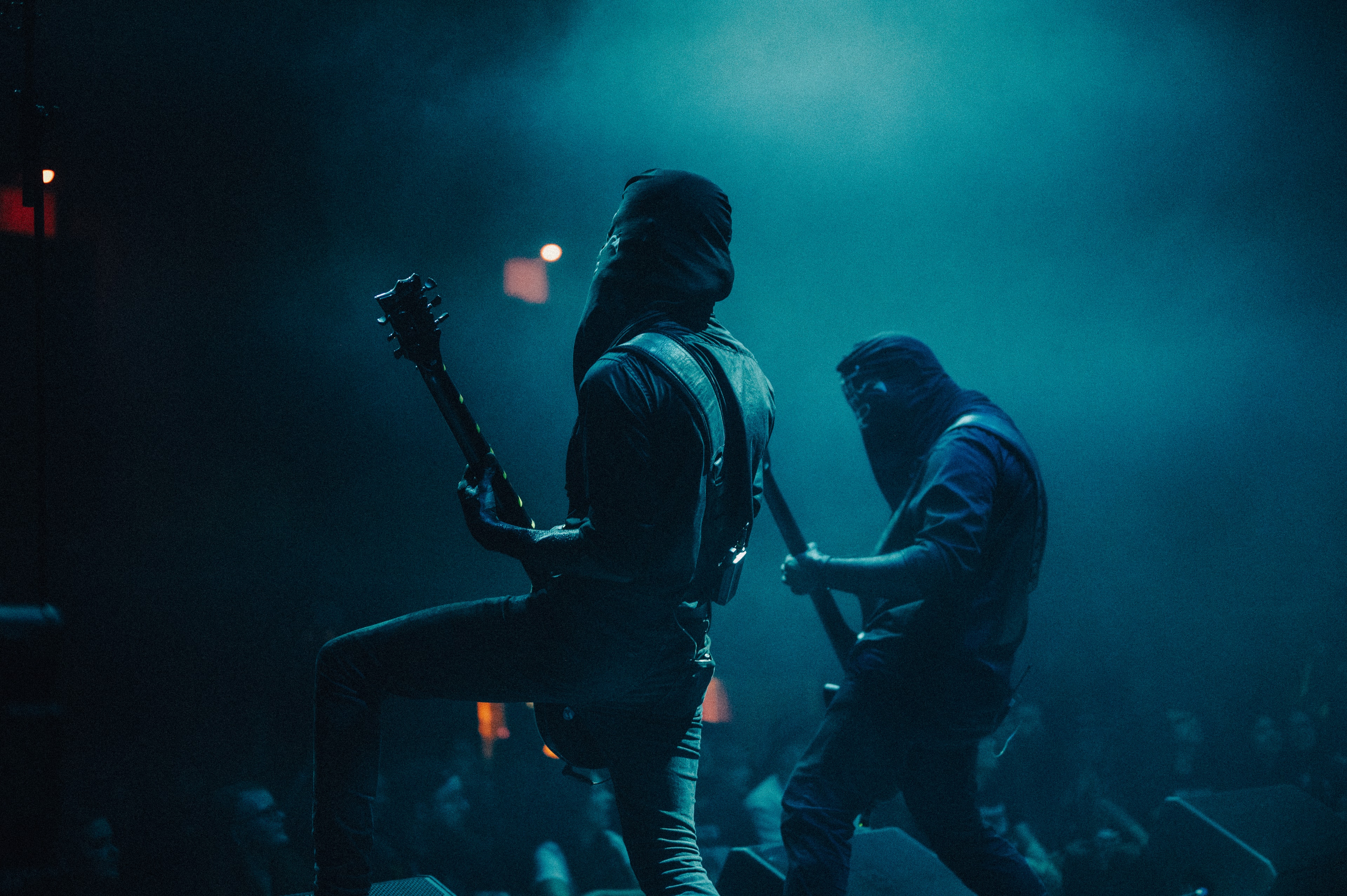 Two hooded, masked guitarists on a smoky stage, front figure with foot propped on a monitor and guitar tilted upward, second musician crouched behind mid-strum, both lit in dim teal-blue haze.