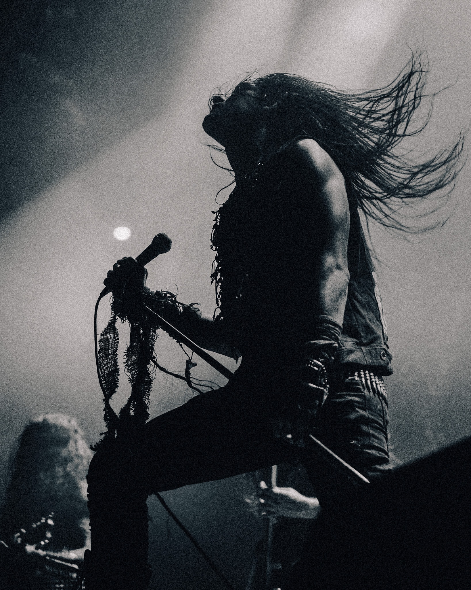 Silhouetted metal vocalist in profile, head tilted back, hair flying upward, one knee raised on stage riser, gripping a mic wrapped in frayed fabric against hazy backlit fog.