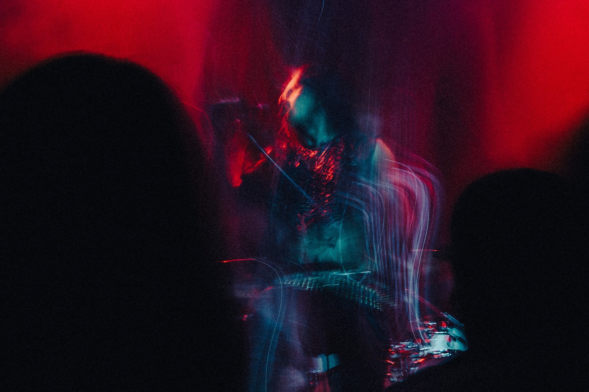 Long-exposure concert photo of a guitarist with head tilted back, red and teal light trails swirling around their torso and instrument, dark audience silhouettes framing left and right.