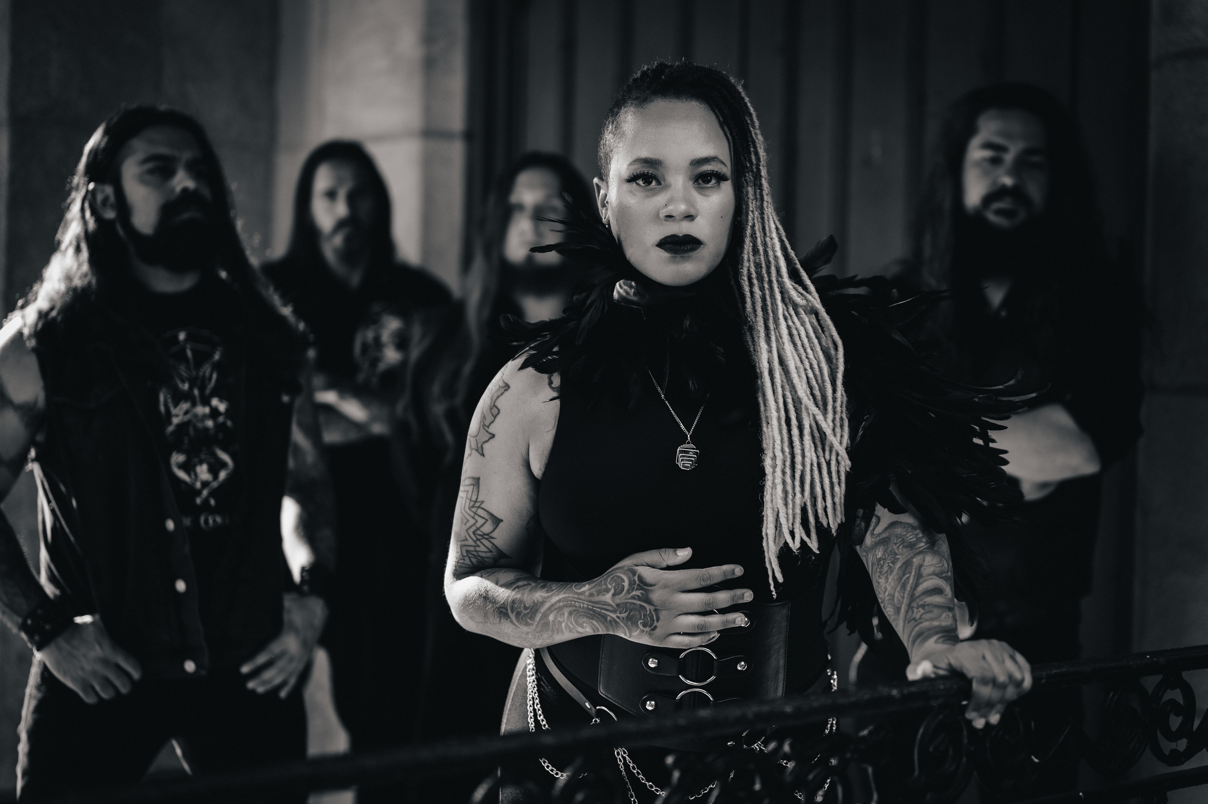 Black-and-white editorial group portrait: a woman with long dreadlocks, dark makeup, and a feathered collar stands in front of four bearded bandmates in a dramatic, gothic-toned session