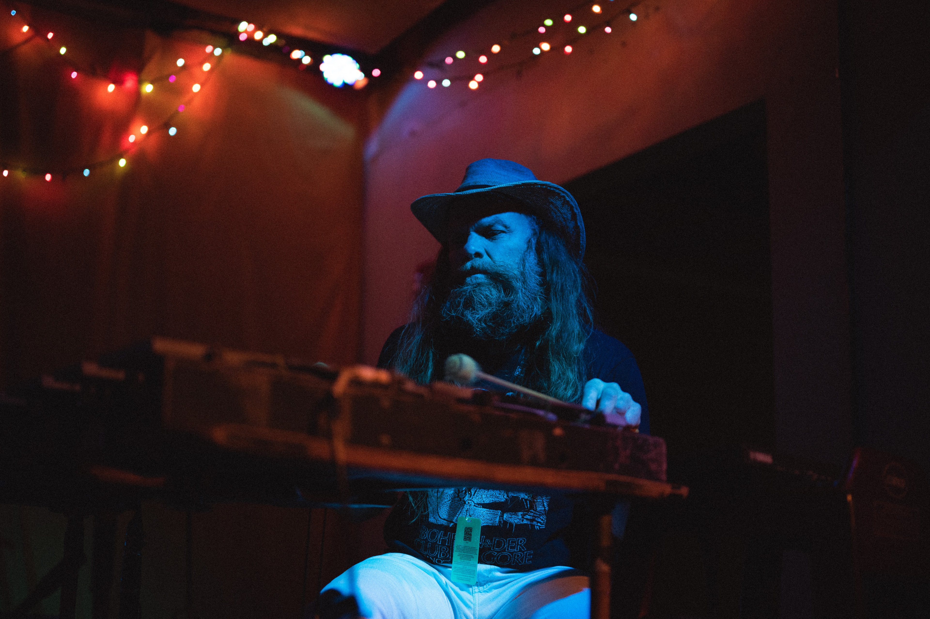Long-haired, bearded man in a fedora hat plays pedal steel guitar with a mallet, blue-lit from below against a dark red wall strung with colorful fairy lights.